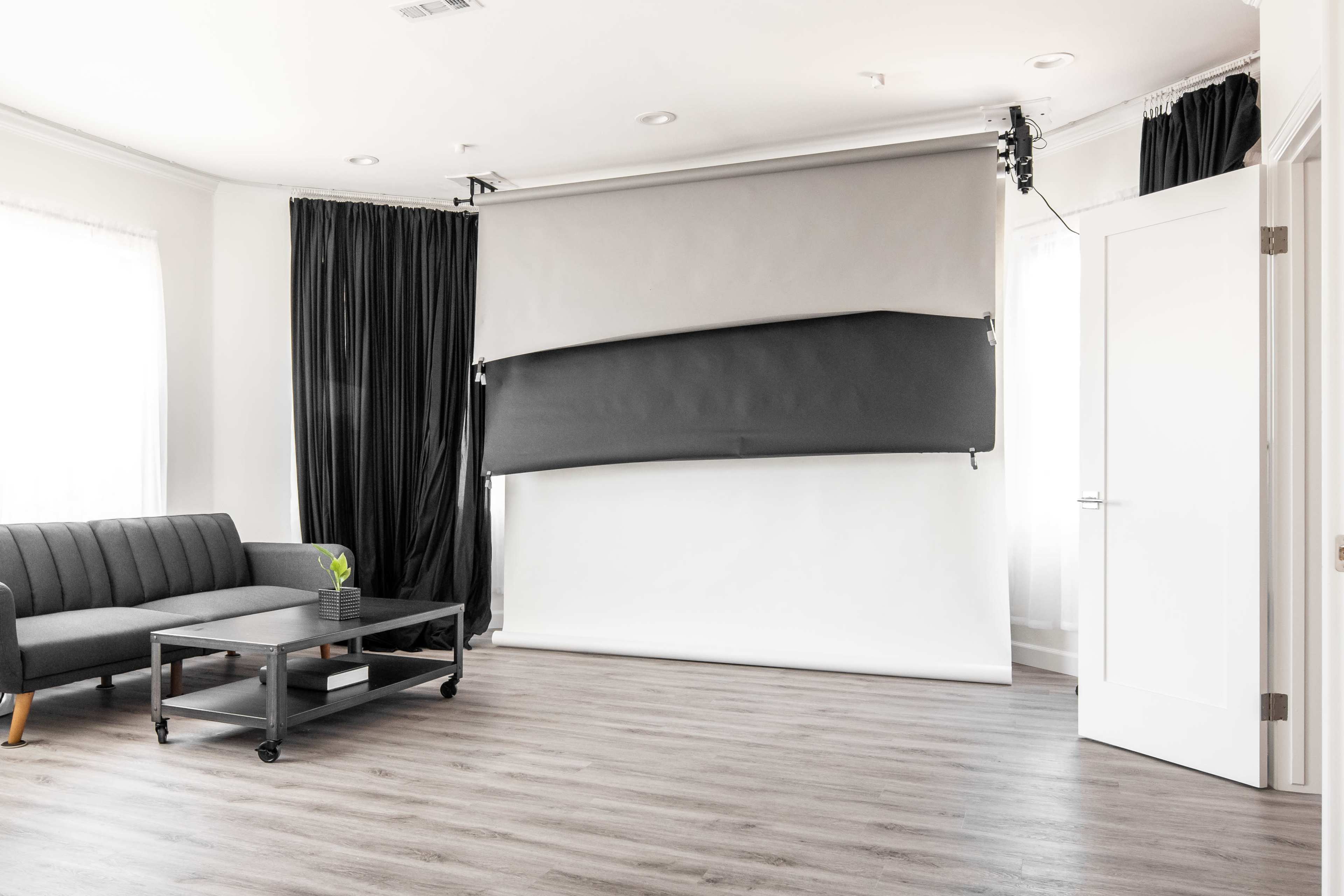 The image shows a minimalist photography studio with a gray backdrop, a black curtain, a gray sofa, and a coffee table on wooden flooring.