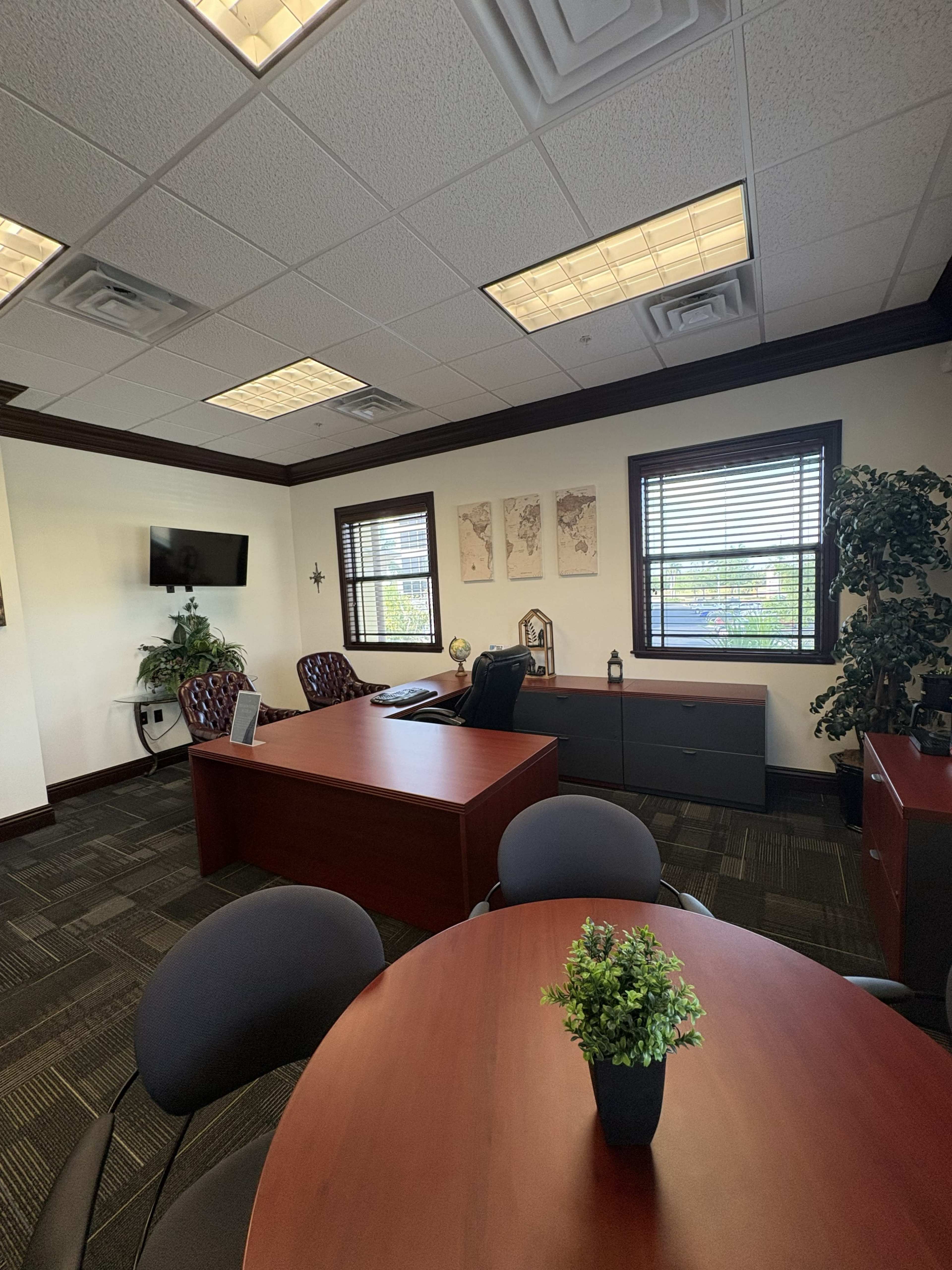 The image shows a modern office space featuring a large desk, a round table with chairs, and decorative elements such as plants and wall art.