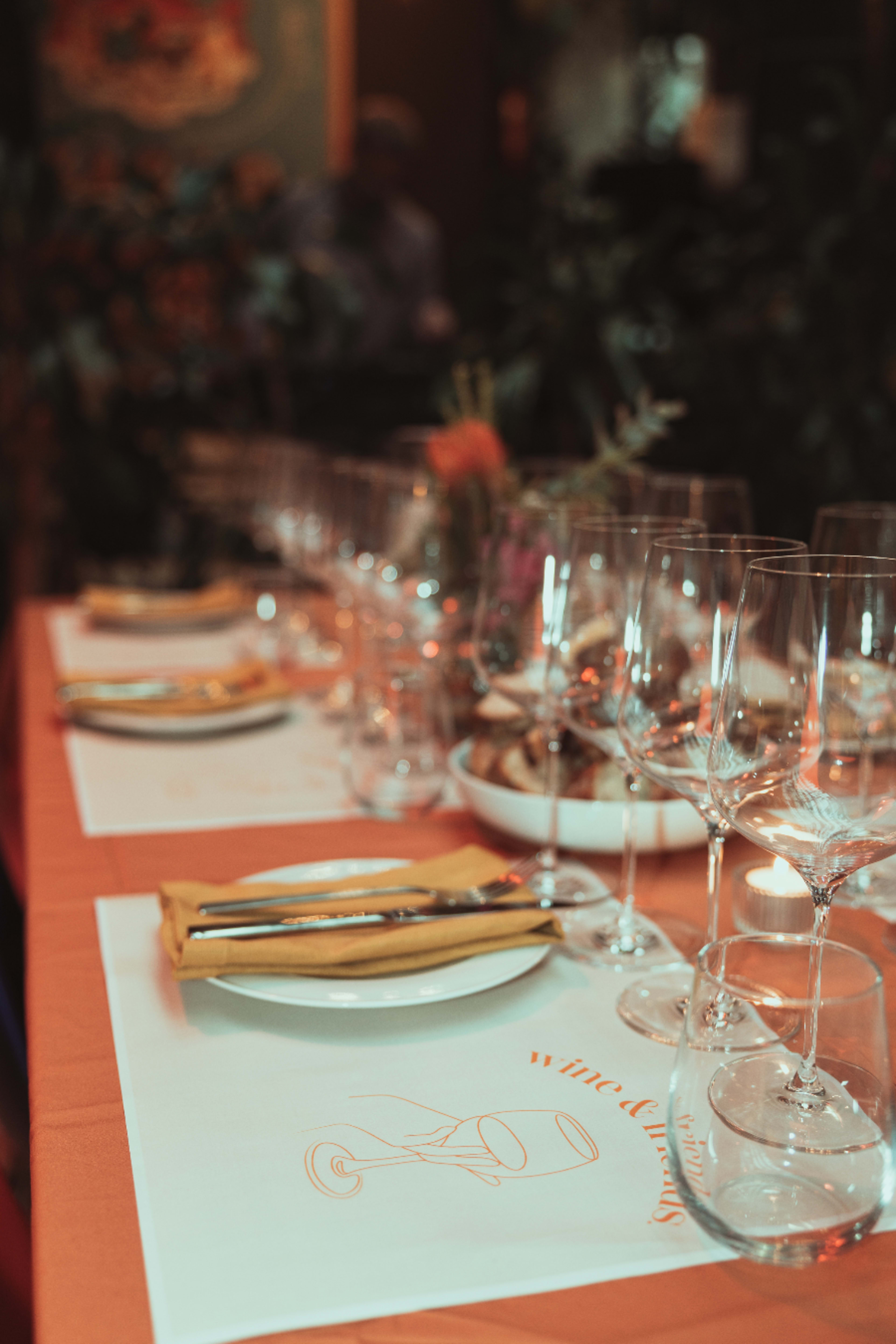 A long dining table is set with plates, wine glasses, and utensils, accompanied by decorative items and a tablecloth featuring wine-themed illustrations.