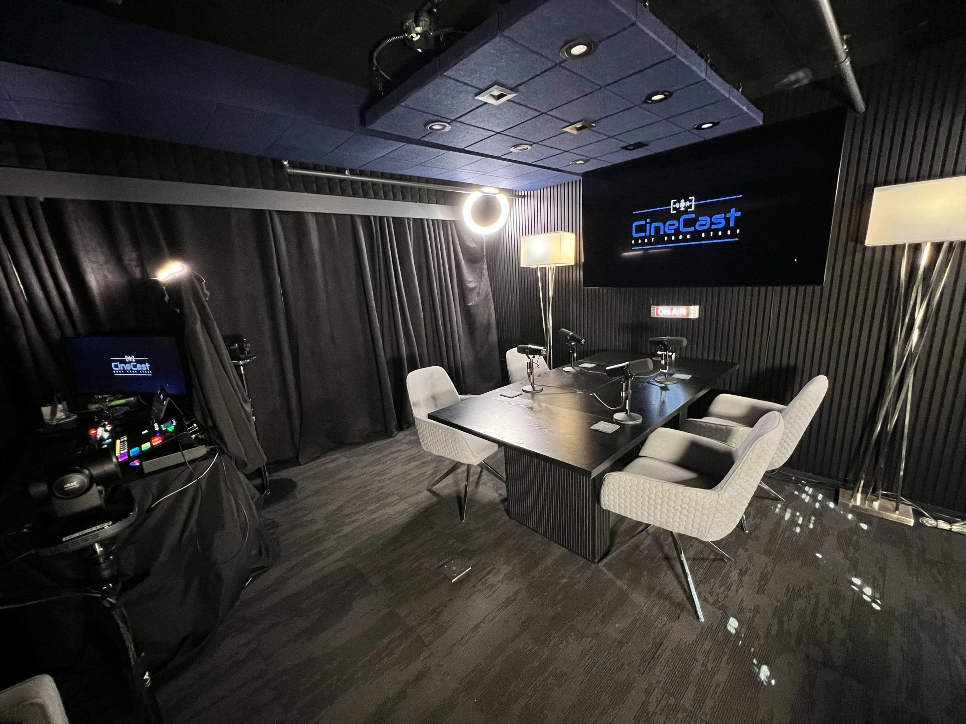 The image shows a modern podcast studio with a large table, two chairs, television screens, and professional lighting equipment.