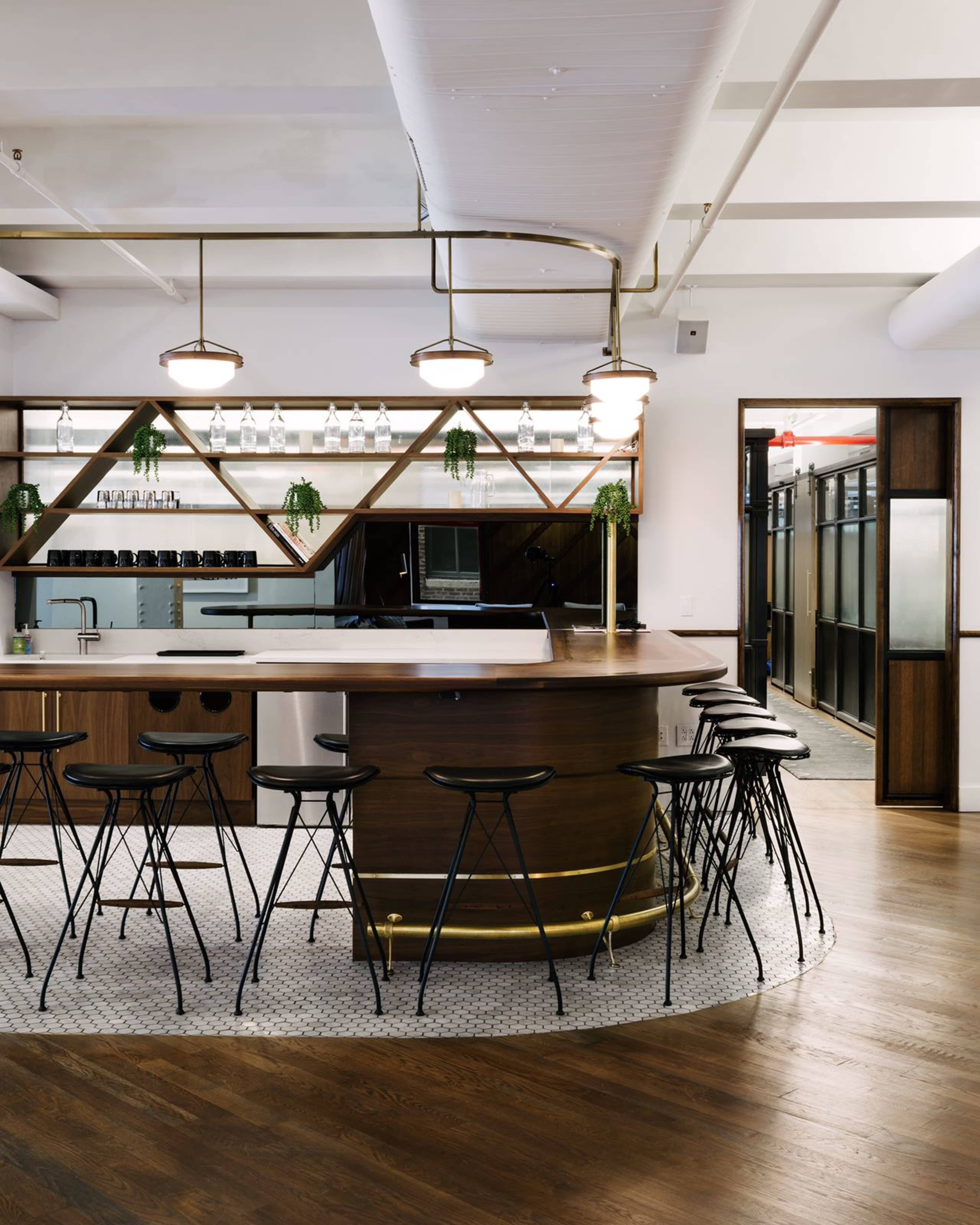 A modern bar area features a circular wooden counter with black stools, surrounded by geometric wooden accents and shelves displaying glassware.