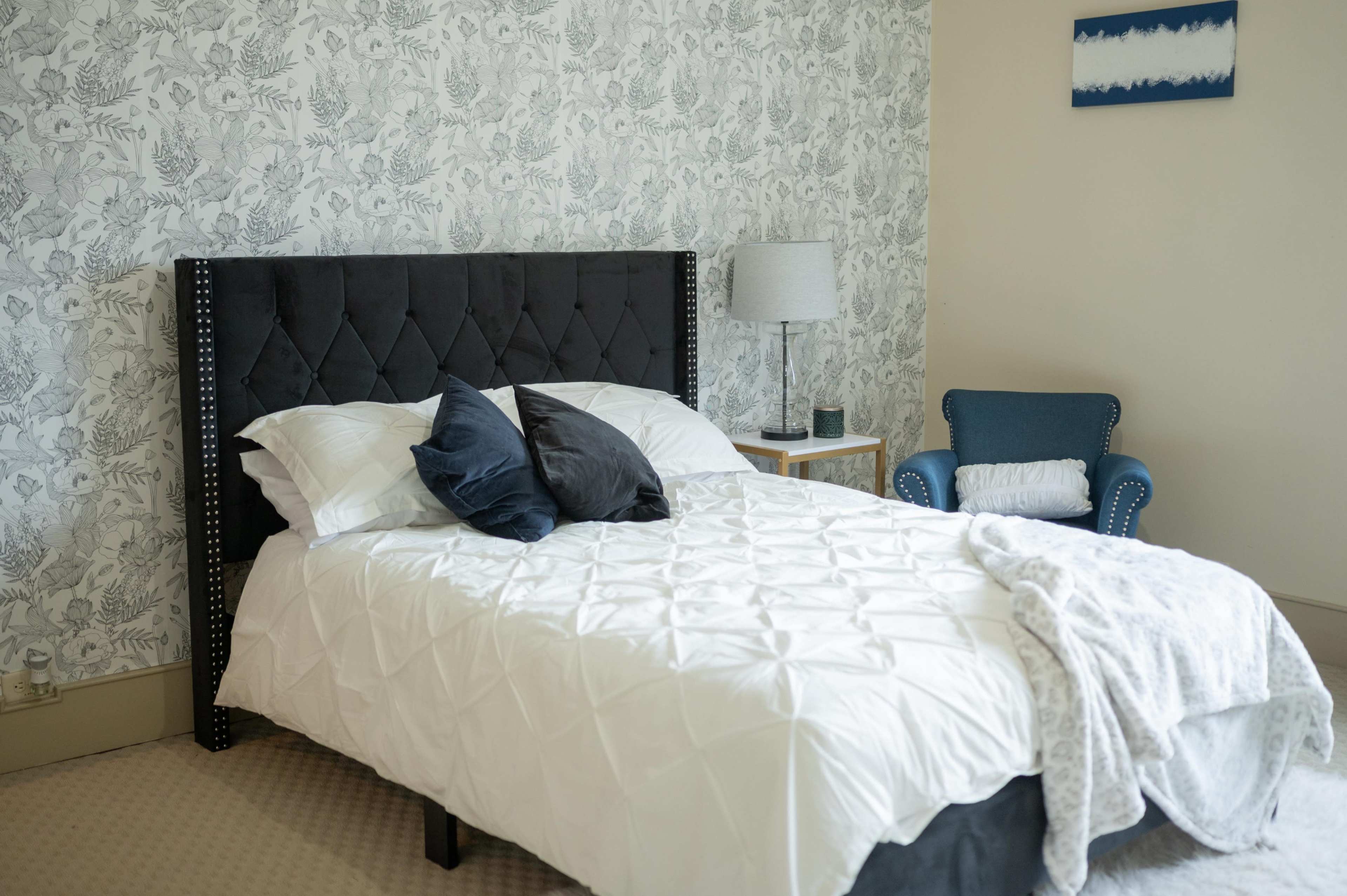 A neatly made bed with a dark upholstered headboard, surrounded by a light floral wallpaper and a blue armchair.