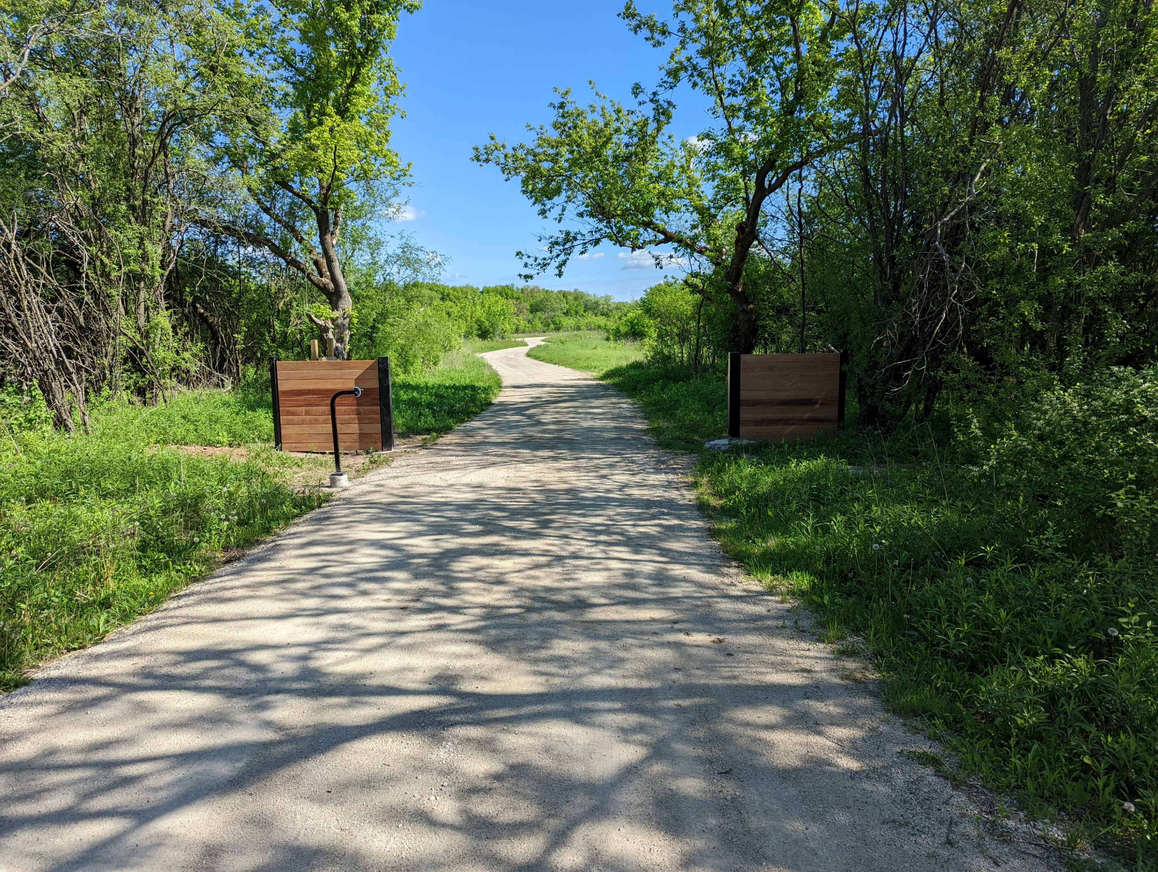 A dirt path winds through a green area, flanked by trees and wooden signs on either side.