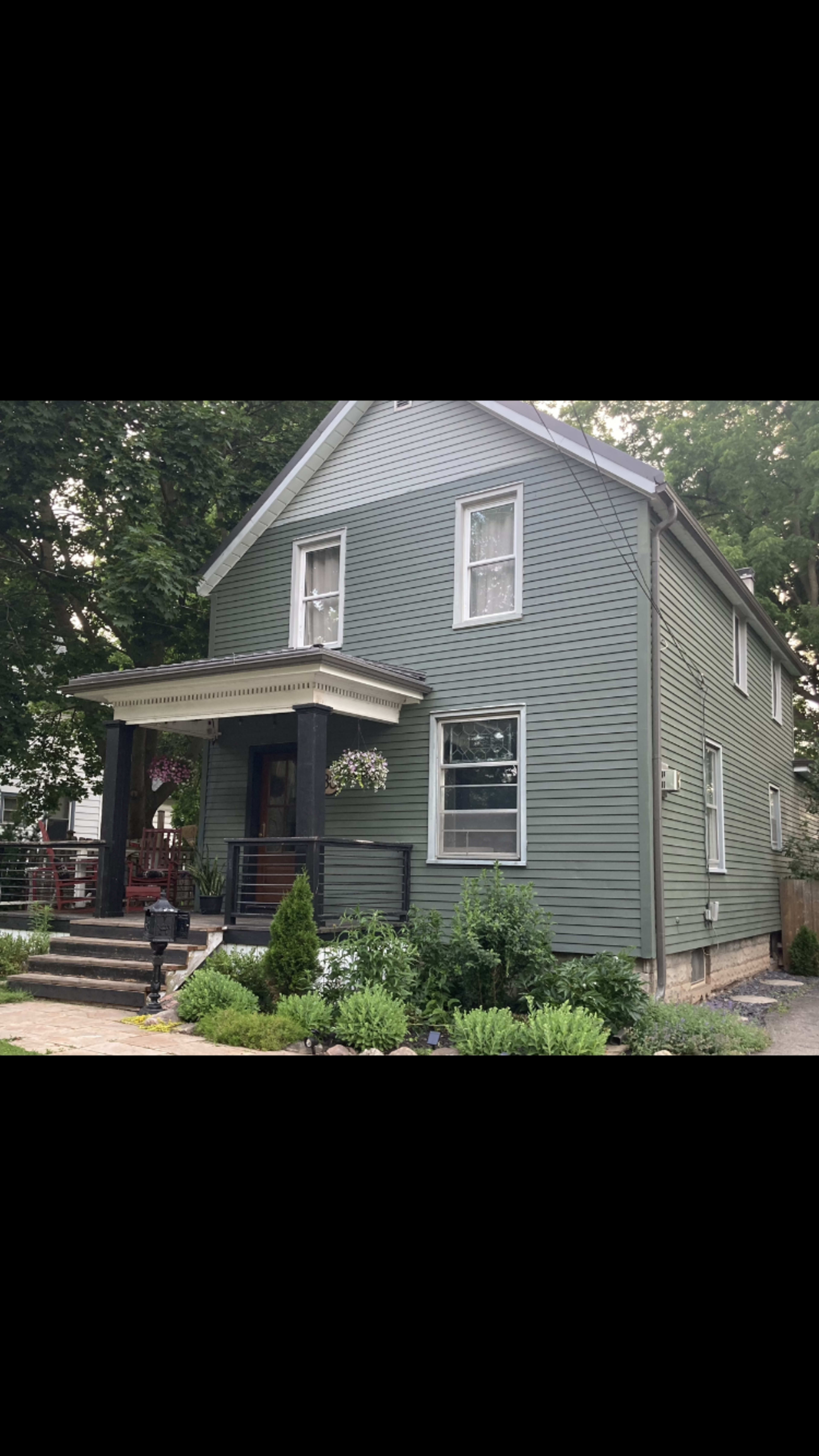 A two-story green house with a front porch, surrounded by shrubs and a landscaped yard.