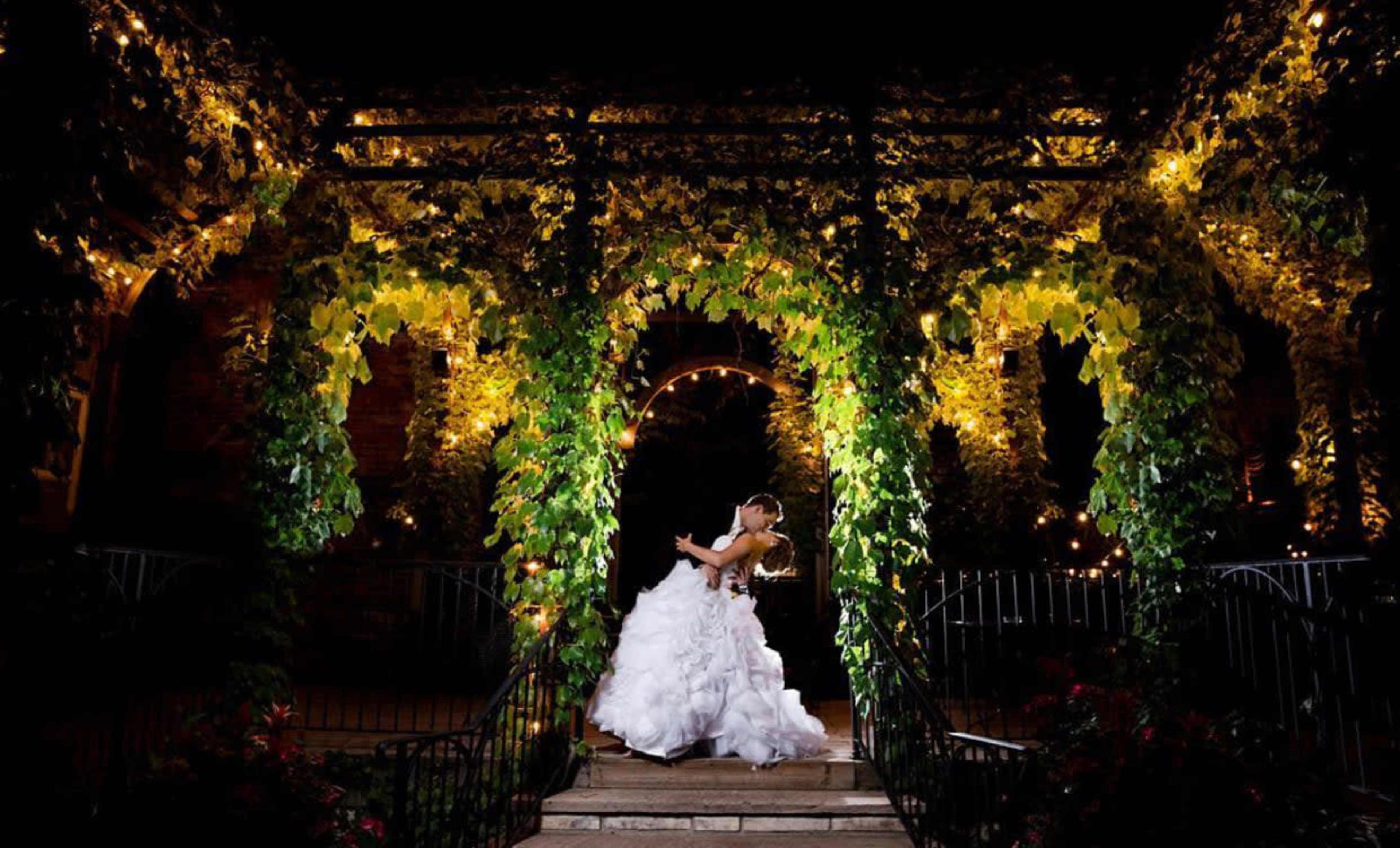 A bride and groom embrace under a vine-covered arch illuminated by warm lights at night.