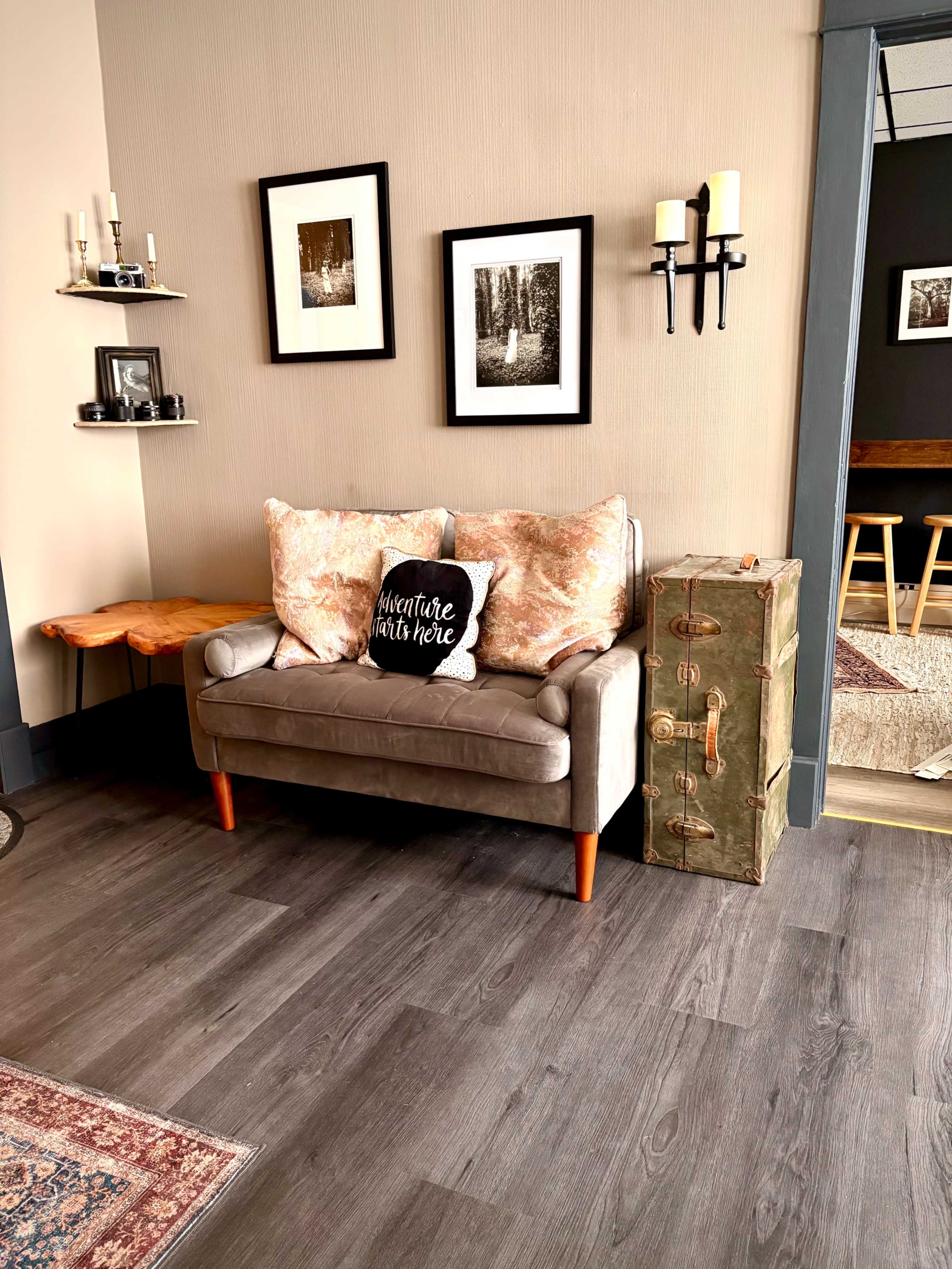 A small, cozy living area features a two-seater sofa with decorative pillows, a wooden side table, and an antique-style trunk beside it, adorned with framed black-and-white photographs on the wall.