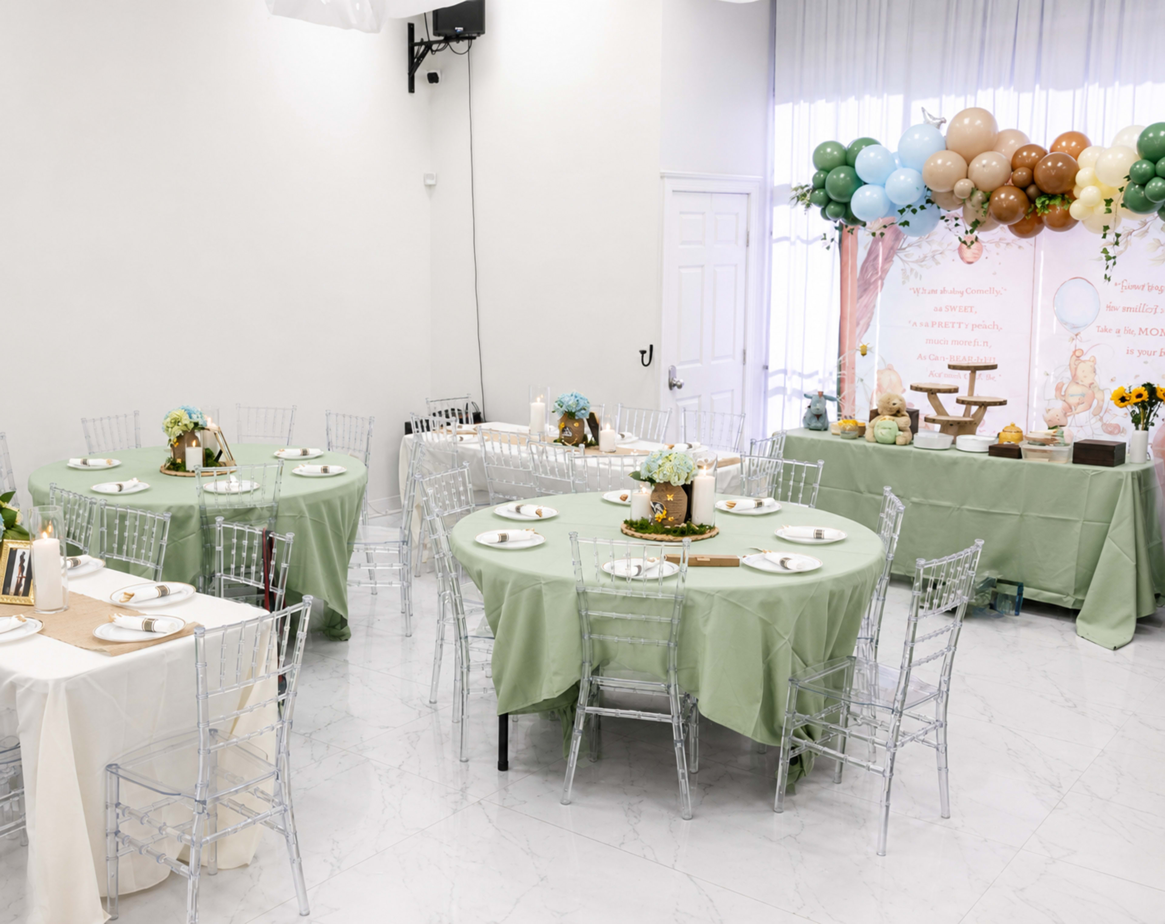 The image shows a decorated event space with round tables dressed in green and neutral tablecloths, each set with plates and floral centerpieces, alongside a backdrop featuring balloons and a decorative banner.