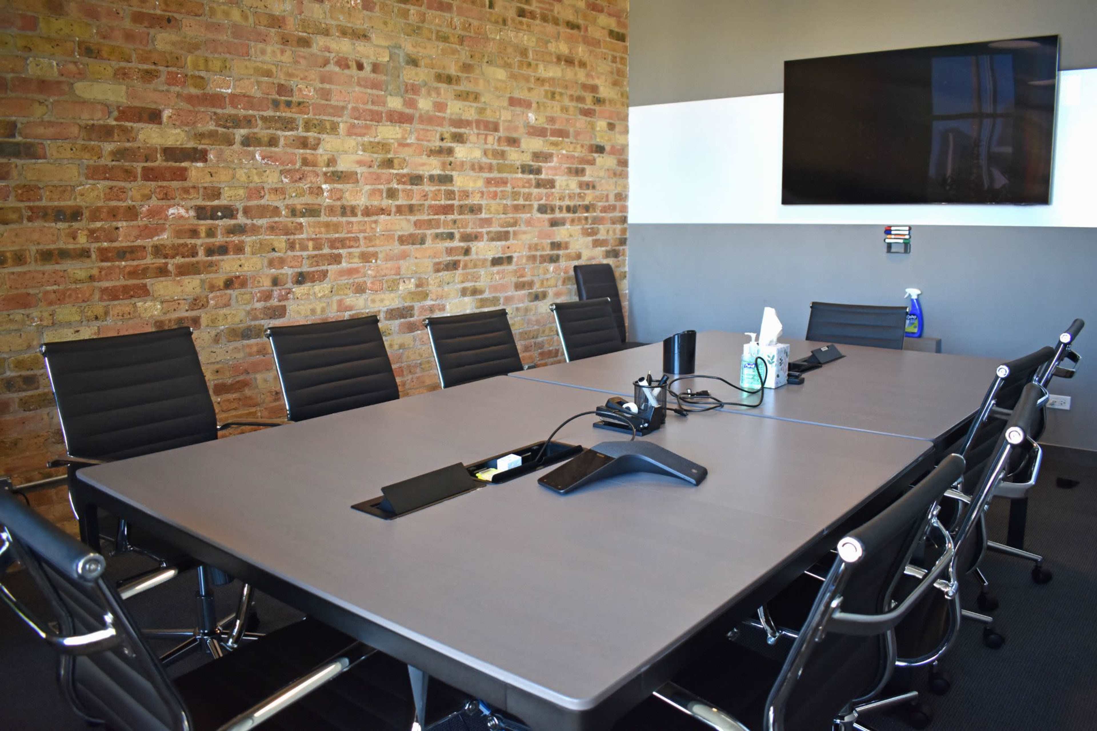 A modern conference room features a large table surrounded by black chairs and a brick wall, with a wall-mounted screen at one end.