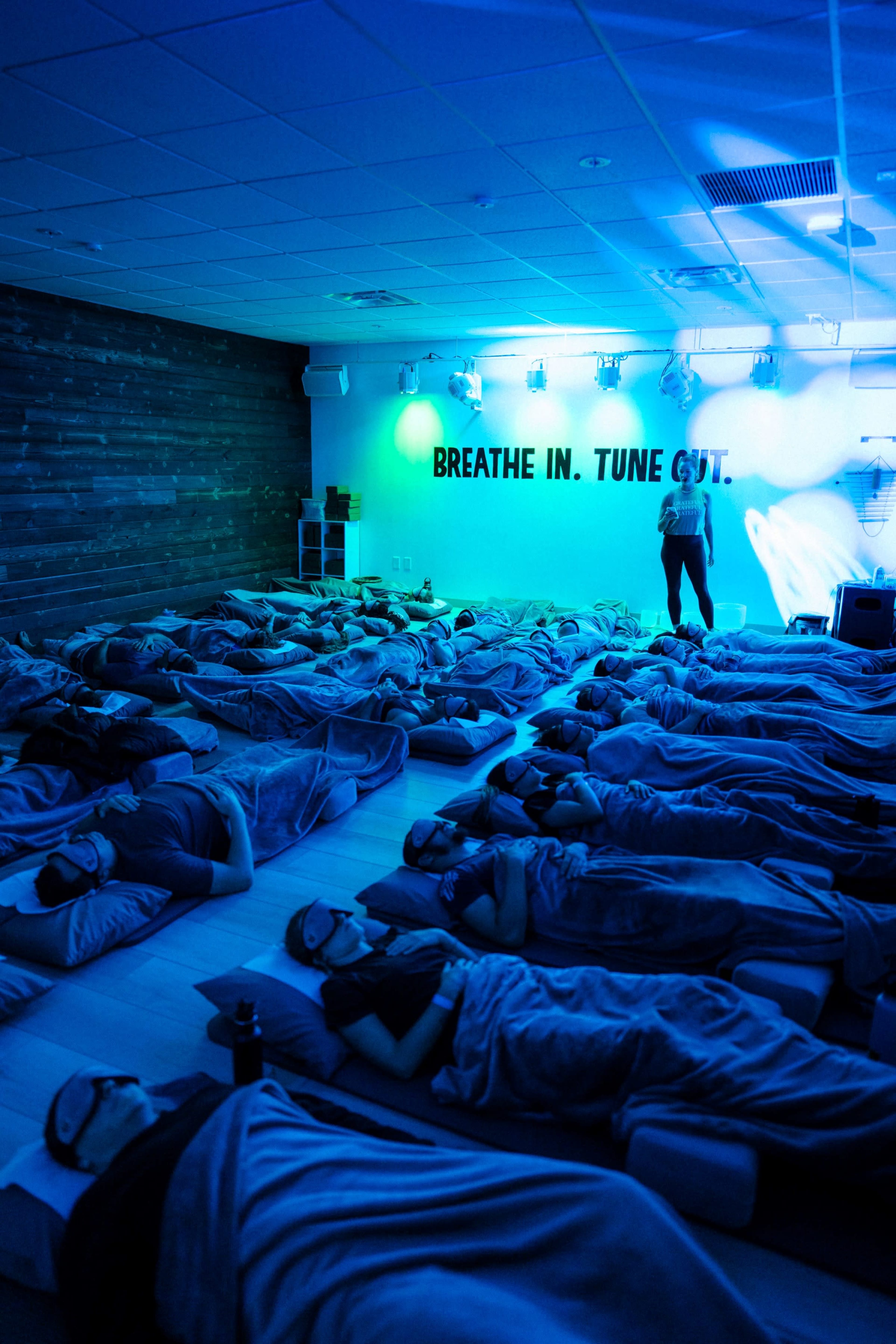 A group of people lies on mats in a dimly lit room, wearing sleep masks and surrounded by blankets, while an instructor stands nearby.