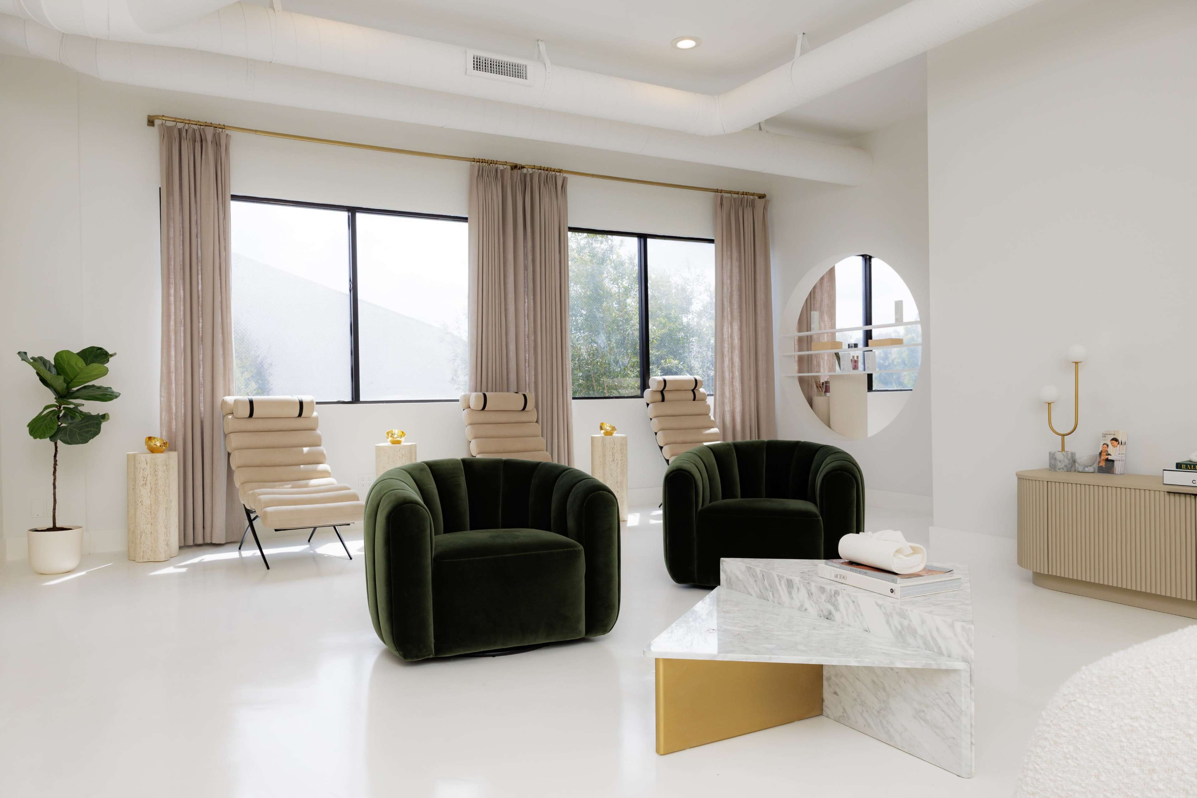 The image shows a modern, minimalist interior featuring two green velvet chairs, a marble coffee table with a gold accent, and large windows dressed with curtains.