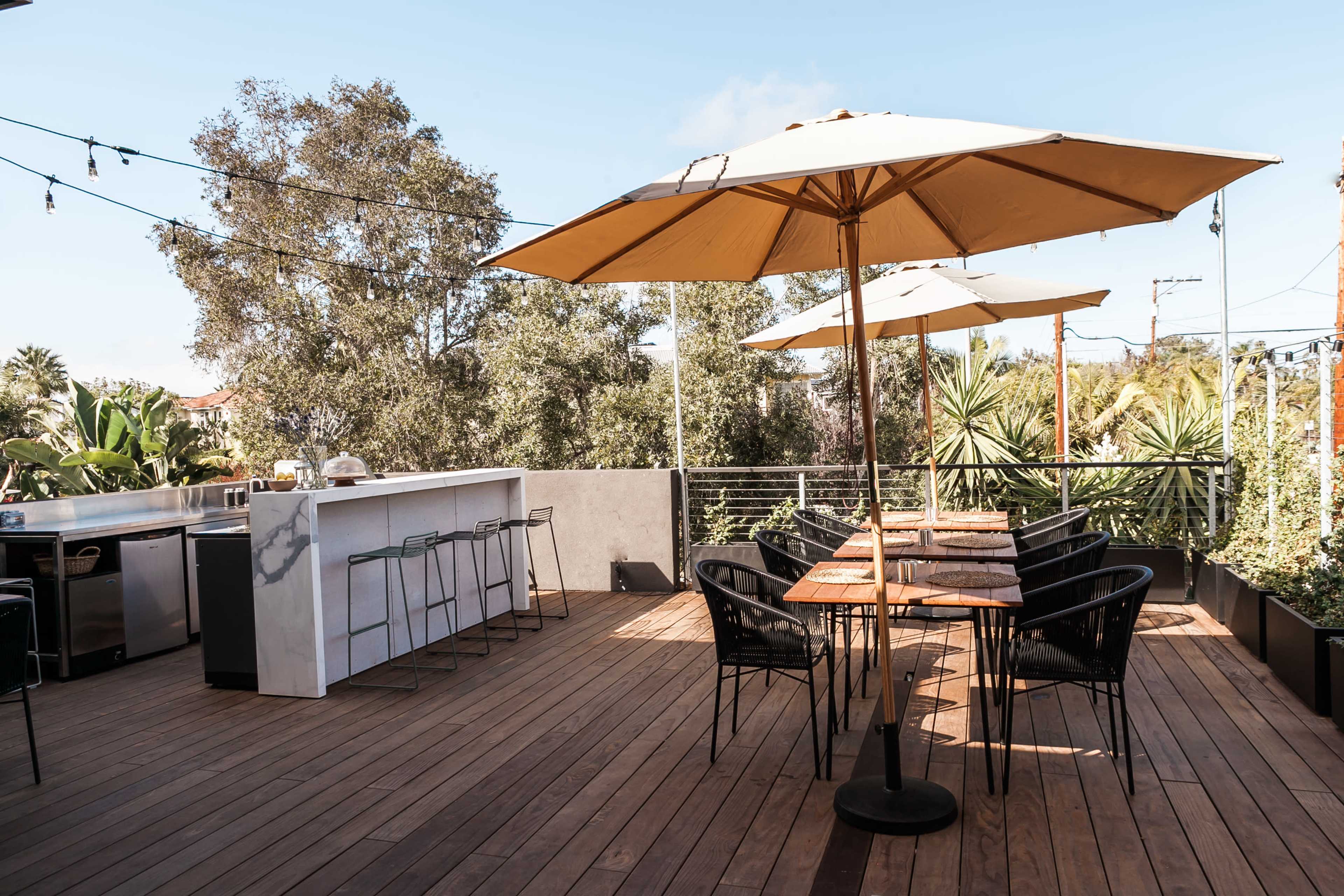 The image shows a modern outdoor patio featuring a bar area, several tables with chairs, and large umbrellas surrounded by greenery.