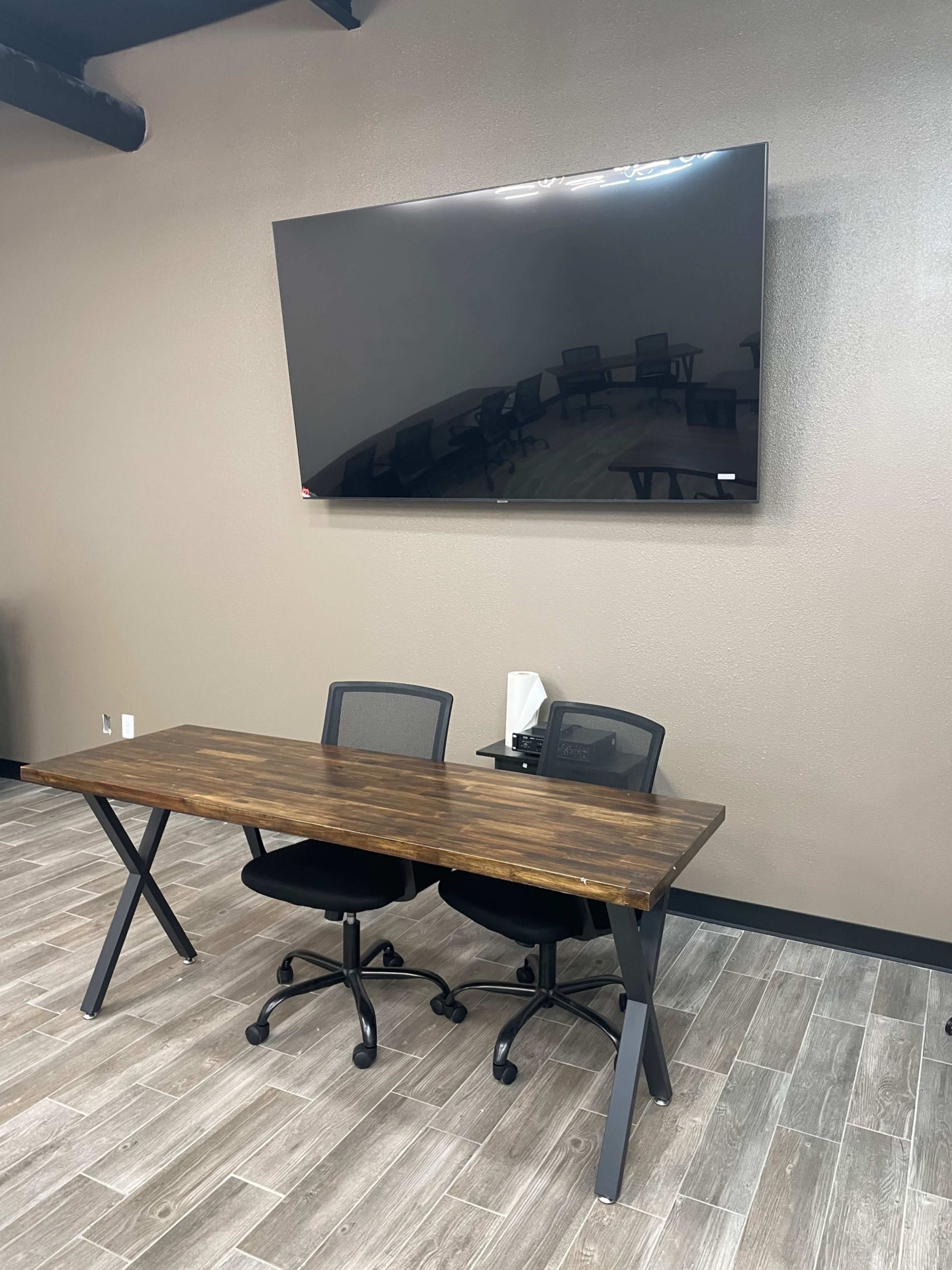 A wooden desk with two black ergonomic chairs positioned in front of a large wall-mounted TV in a modern office space.