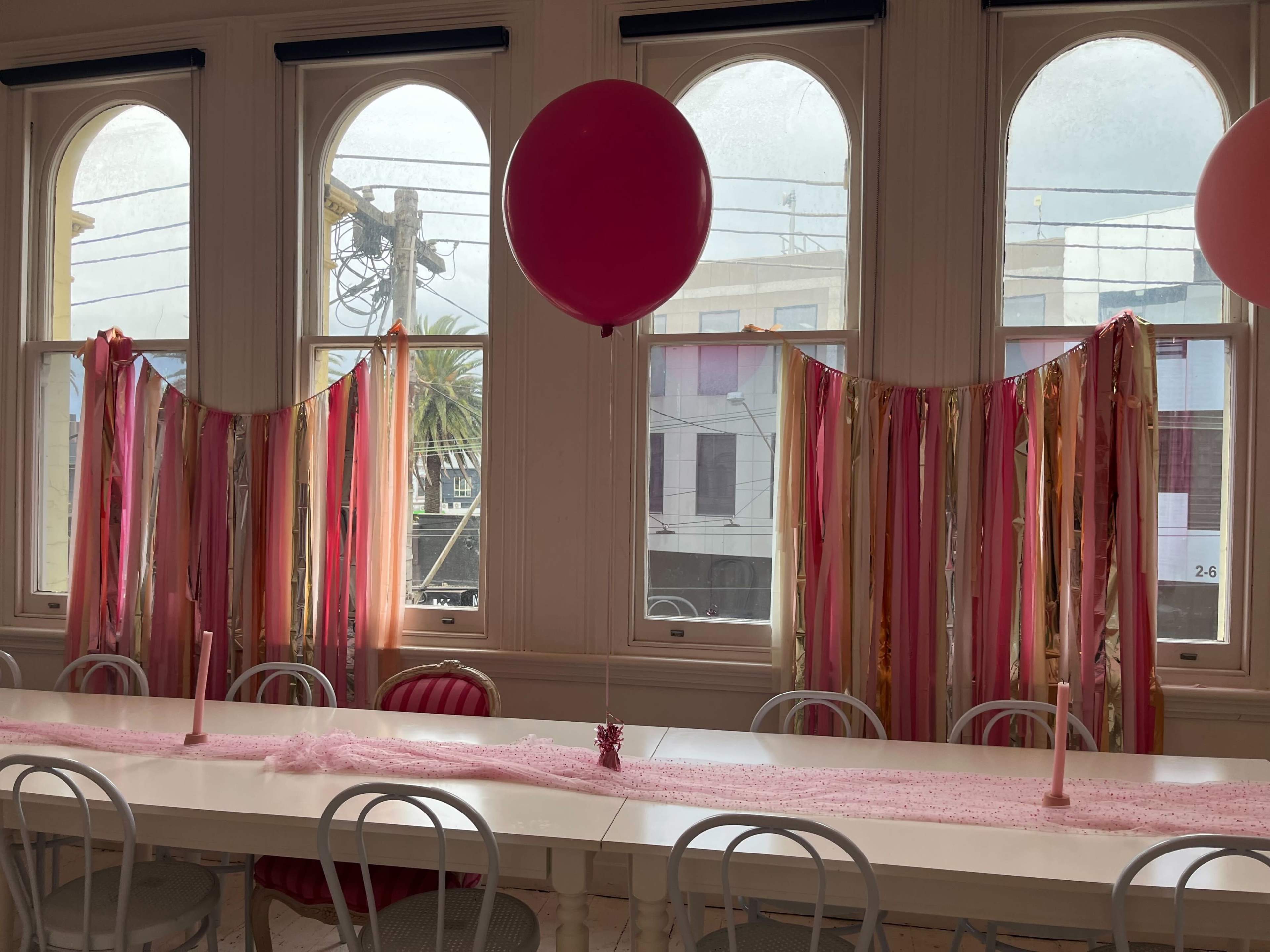 A long table is set up in a brightly lit room with large windows, decorated with pink and gold streamers, candles, and a large pink balloon.