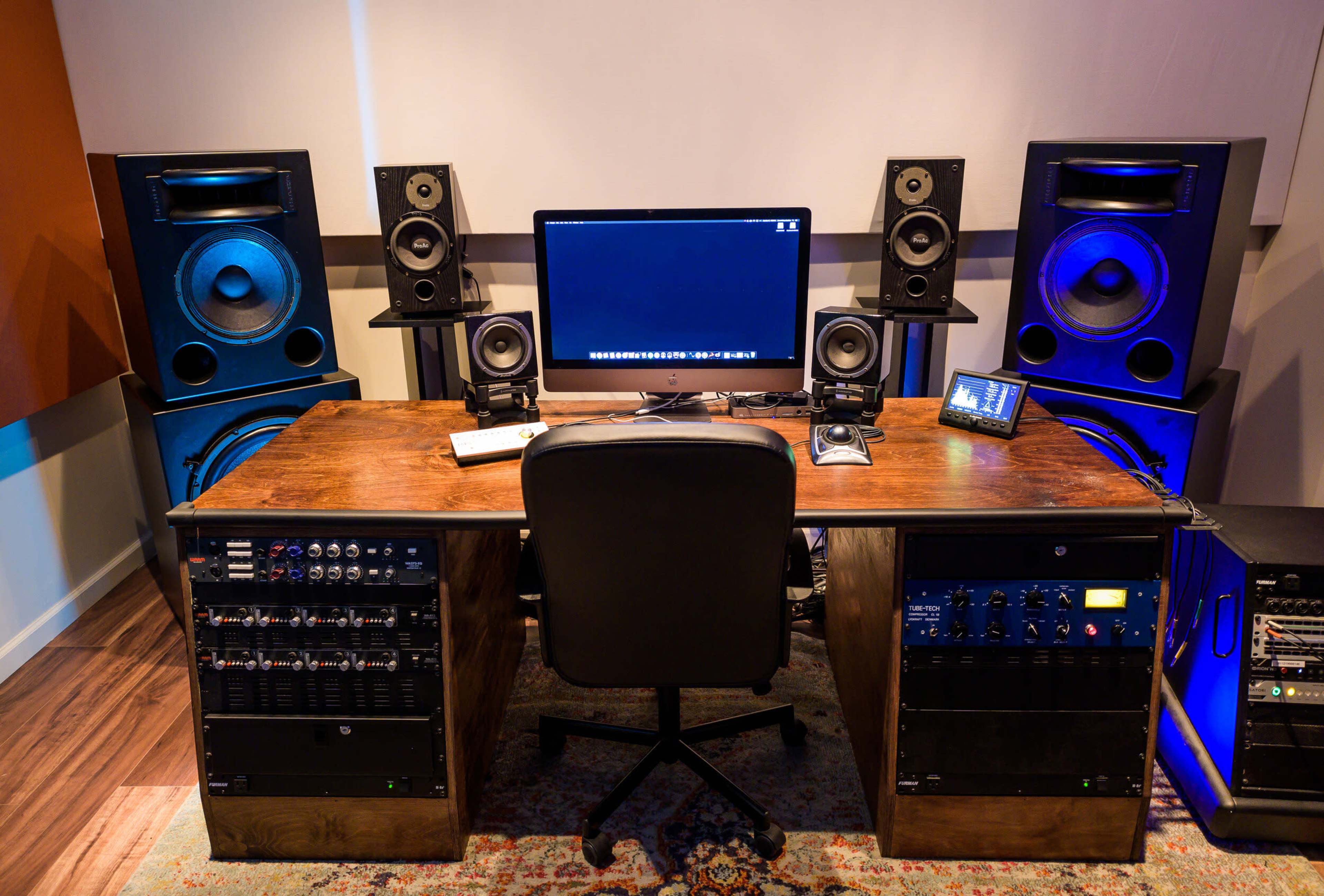 A modern audio production studio features a wooden desk with a computer and various music equipment, flanked by large speakers on either side.