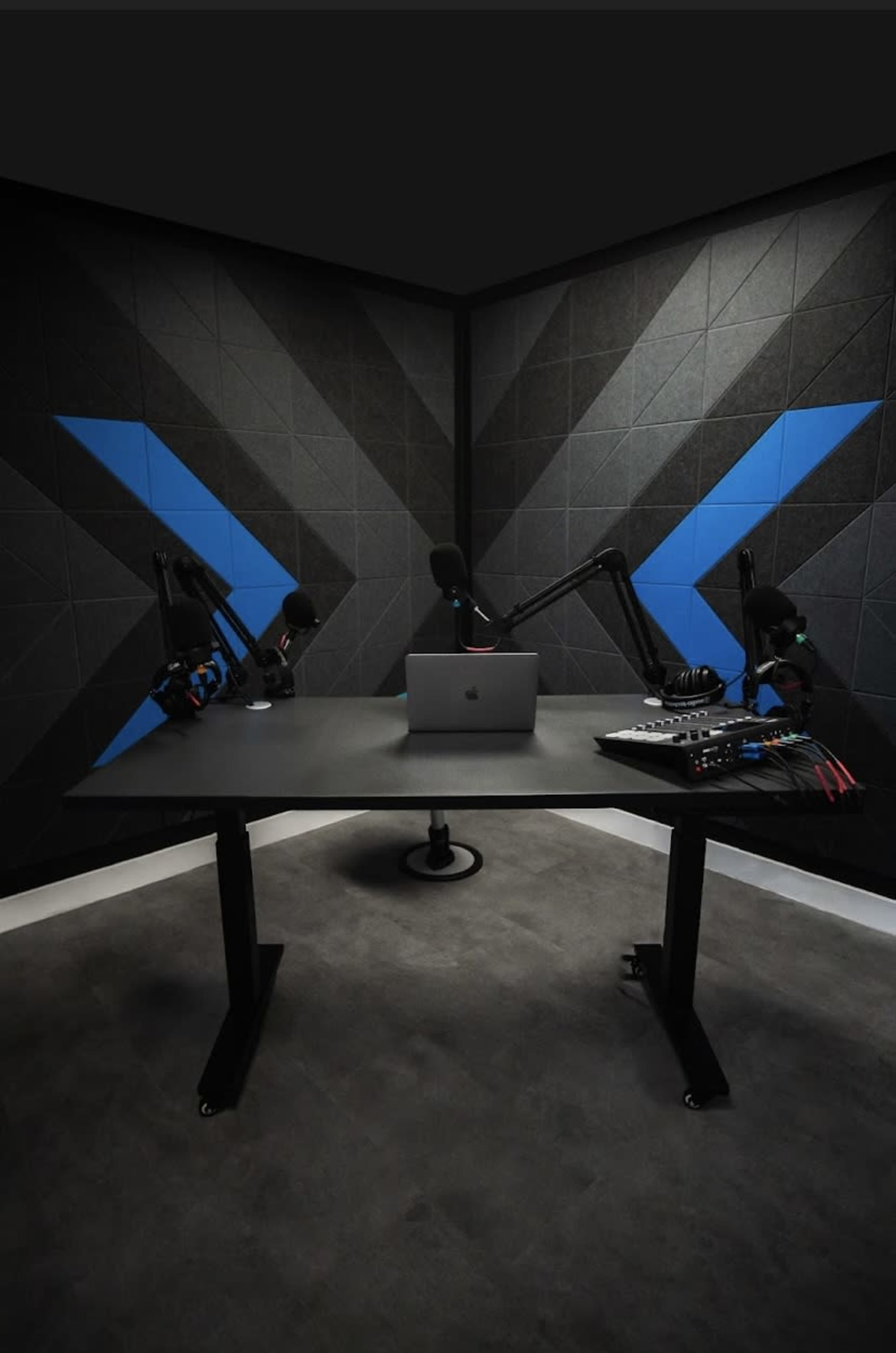 A podcast studio with a desk, a laptop, audio equipment, and two microphone setups against a backdrop of textured gray walls featuring blue triangular patterns.