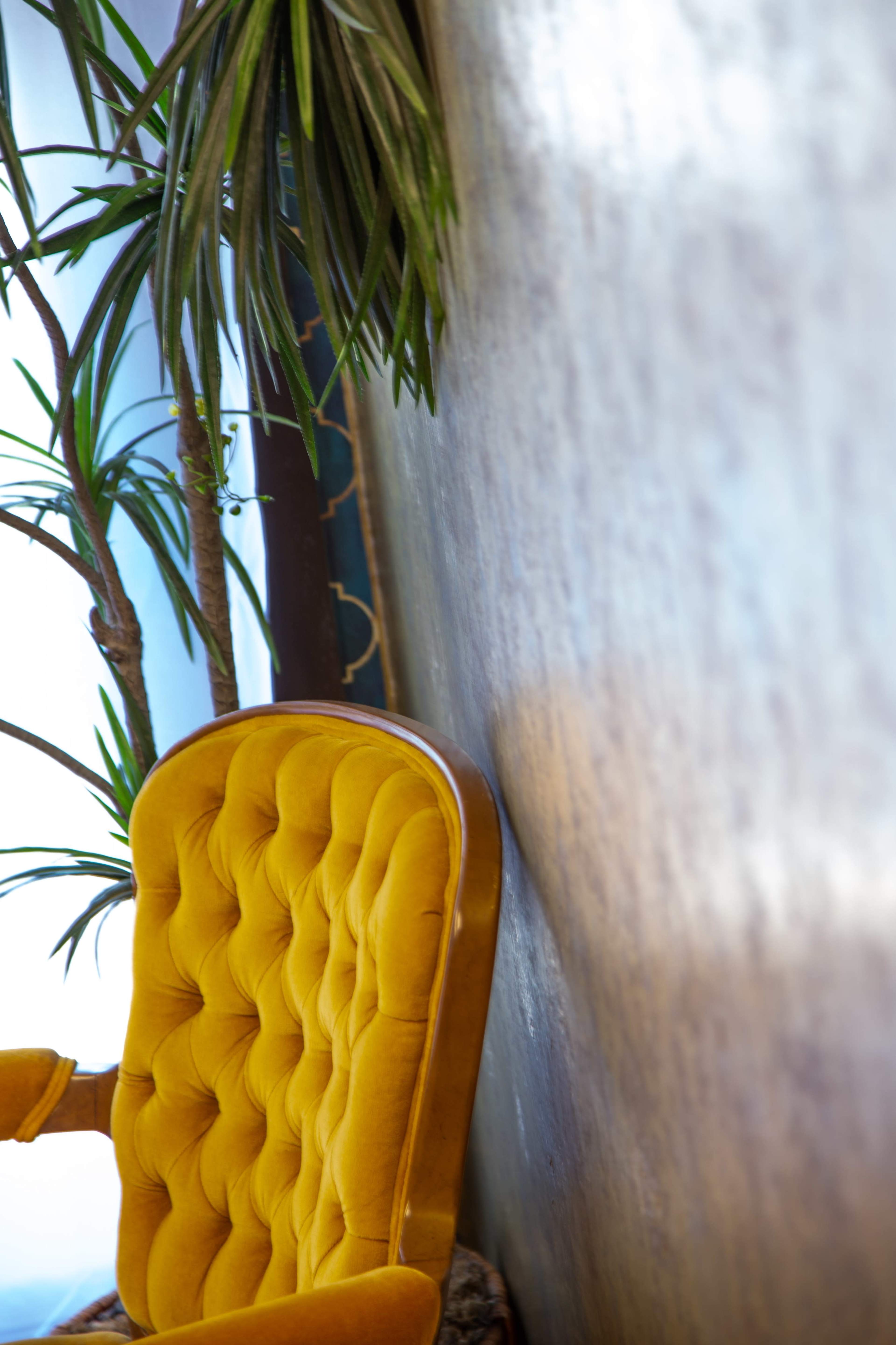 A tufted yellow chair leans against a textured wall with potted plants nearby.