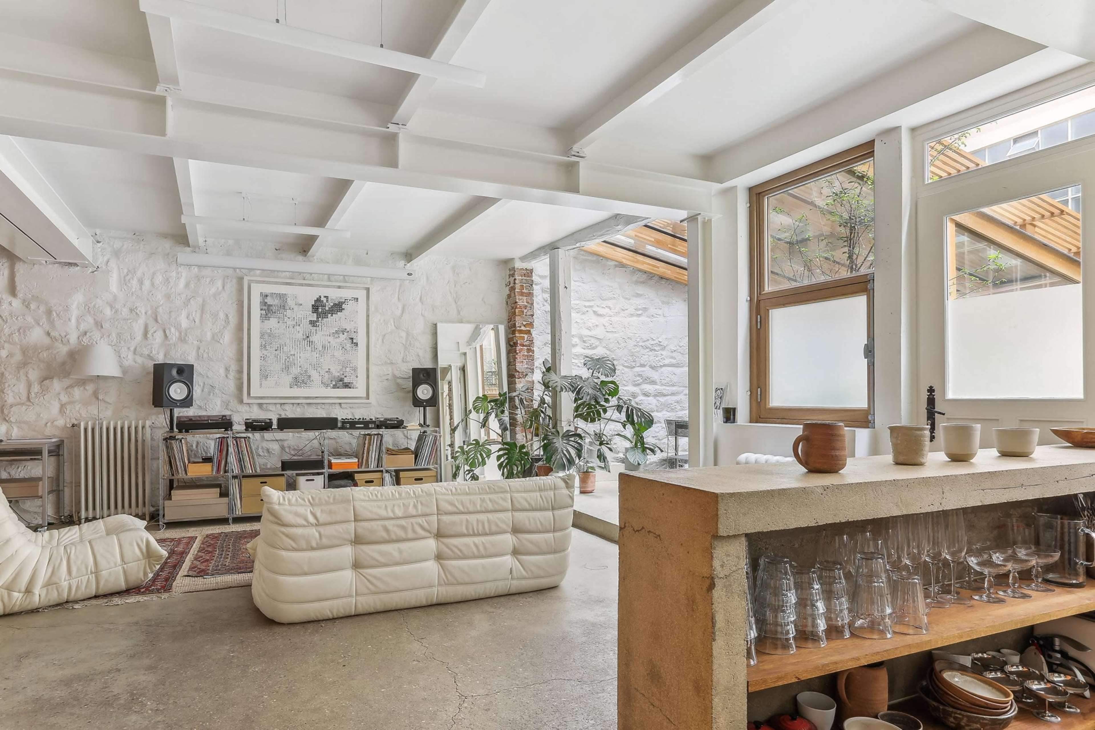 The image shows a modern, open-concept living space featuring a unique white sofa, a wooden kitchen counter with glassware, and large windows that allow natural light to fill the room.