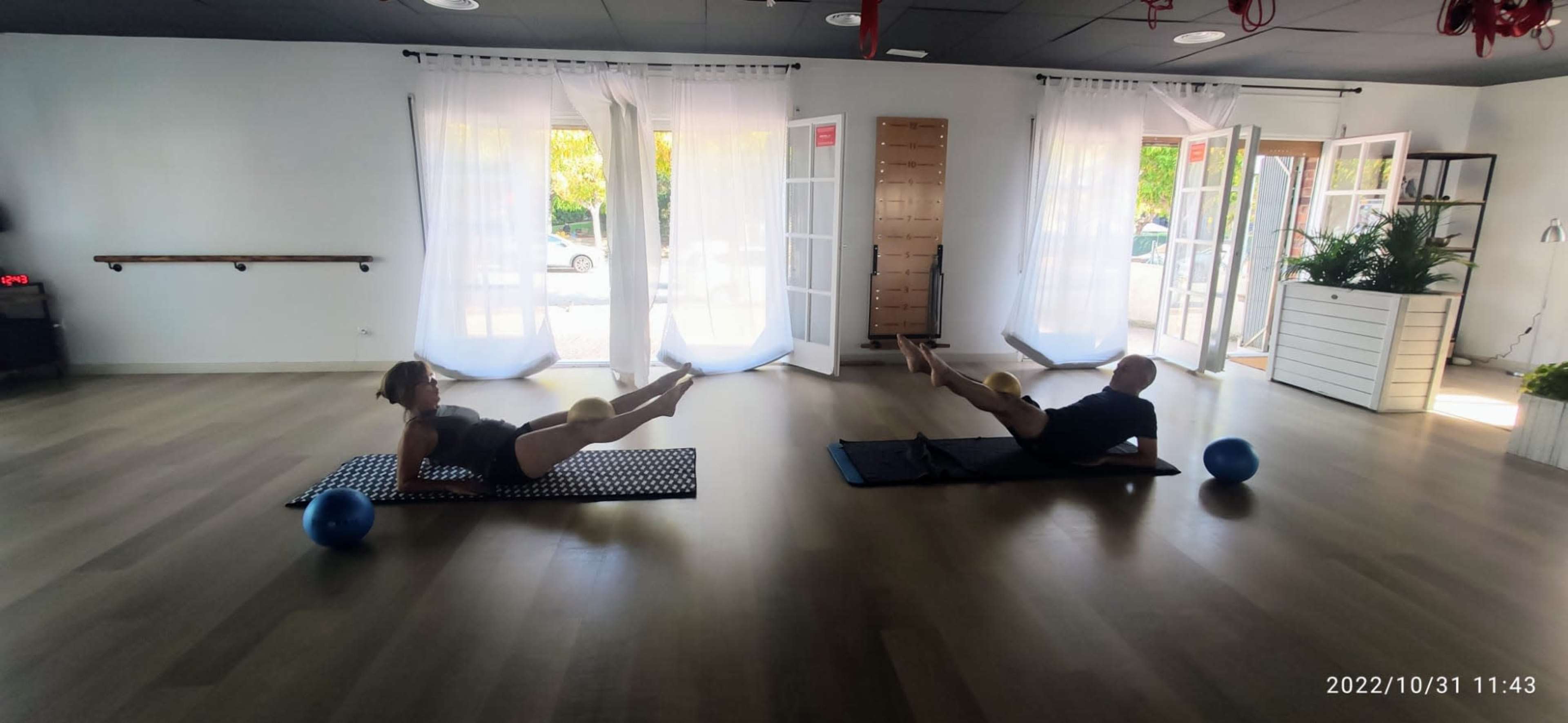 Two individuals are performing leg exercises on mats in a bright, spacious fitness studio.