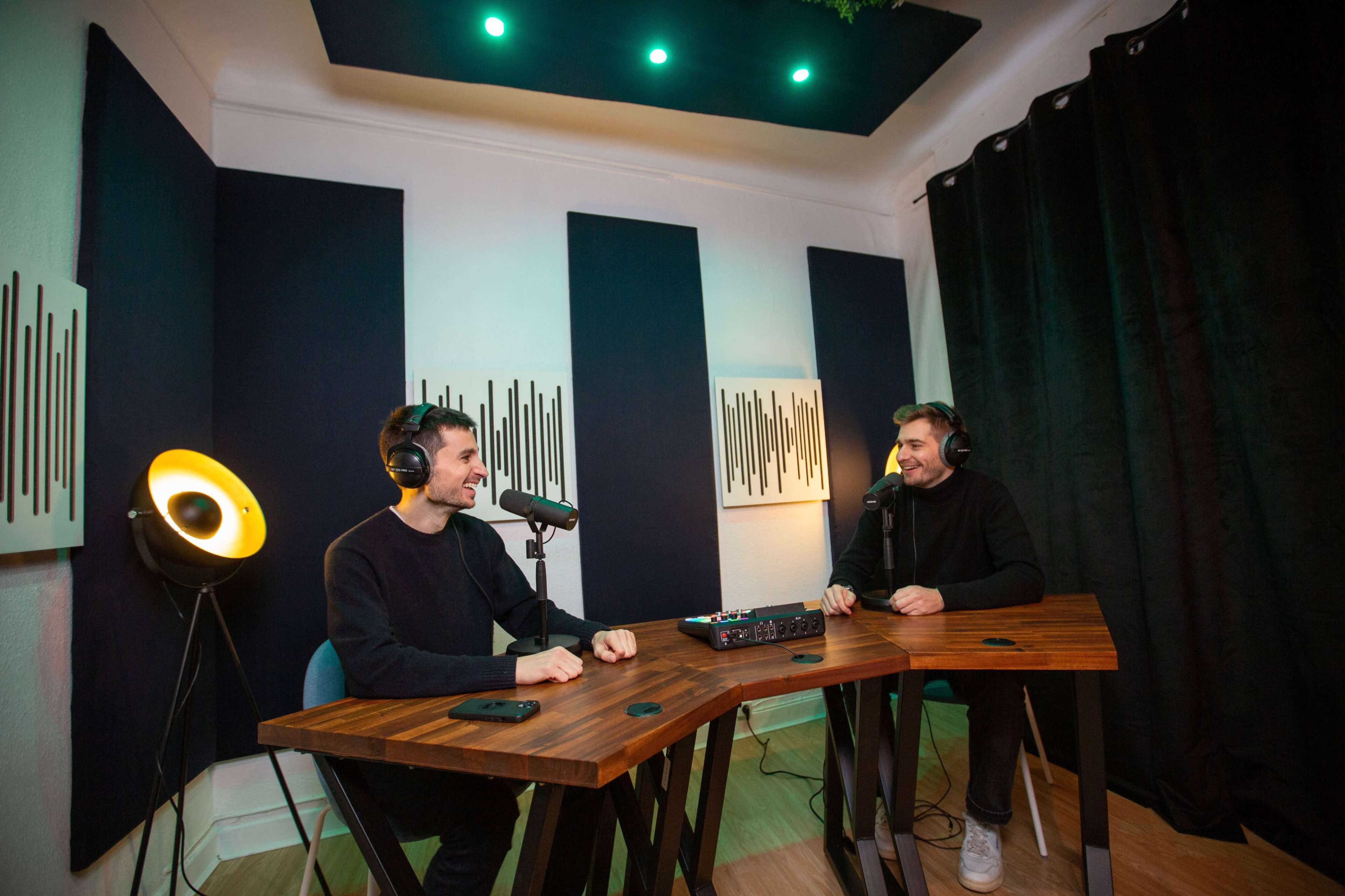 Two men sit at a wooden table with microphones in front of them in a recording studio with soundproofing panels and ambient lighting.