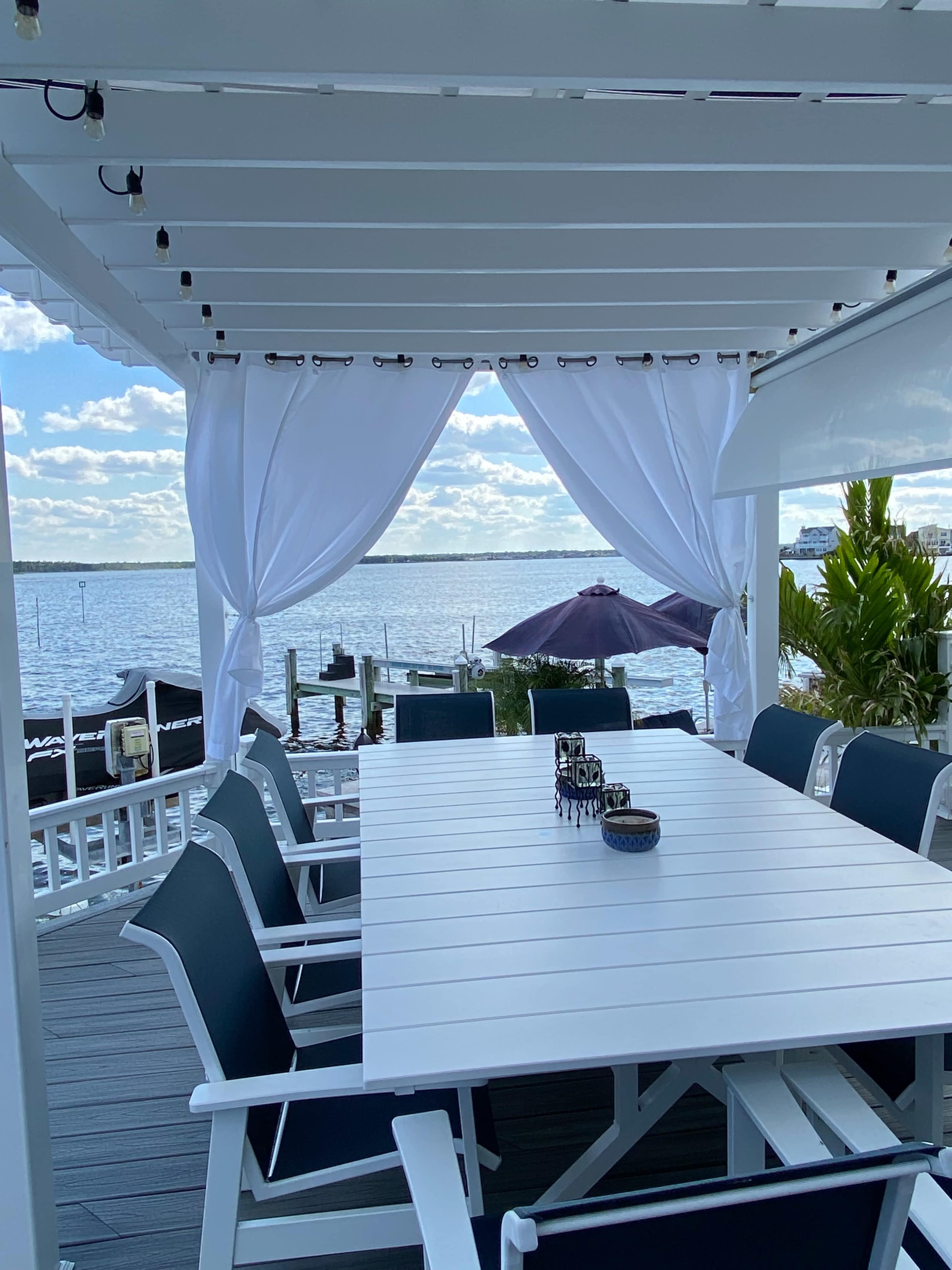 A white table with black chairs is set on a deck overlooking a body of water, framed by flowing white curtains.