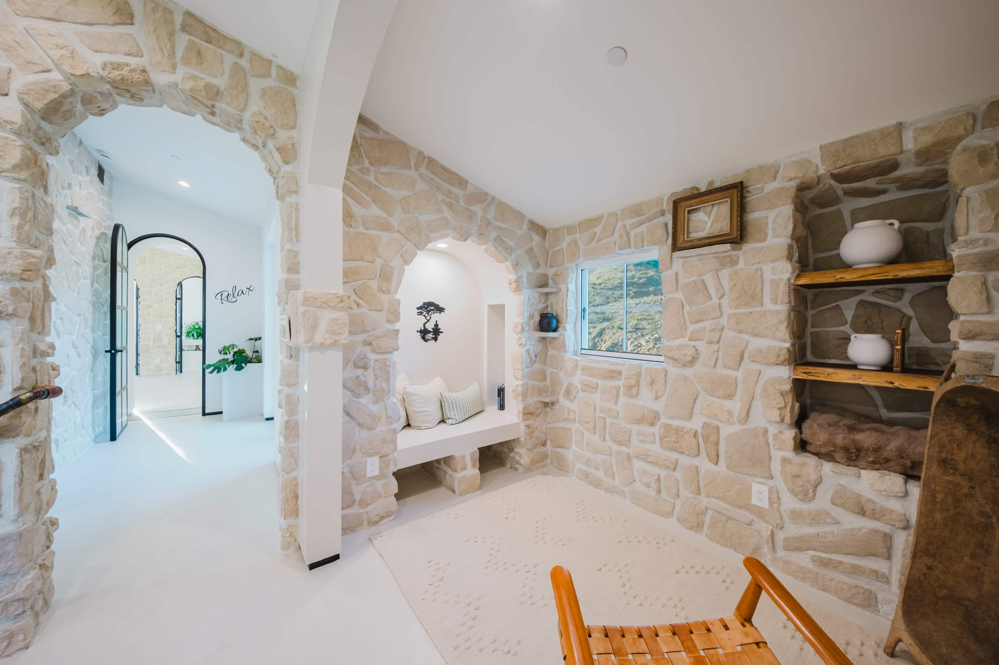 The image shows a light-filled interior with textured stone walls, a cozy seating nook, and a view of a window framed by additional stonework.