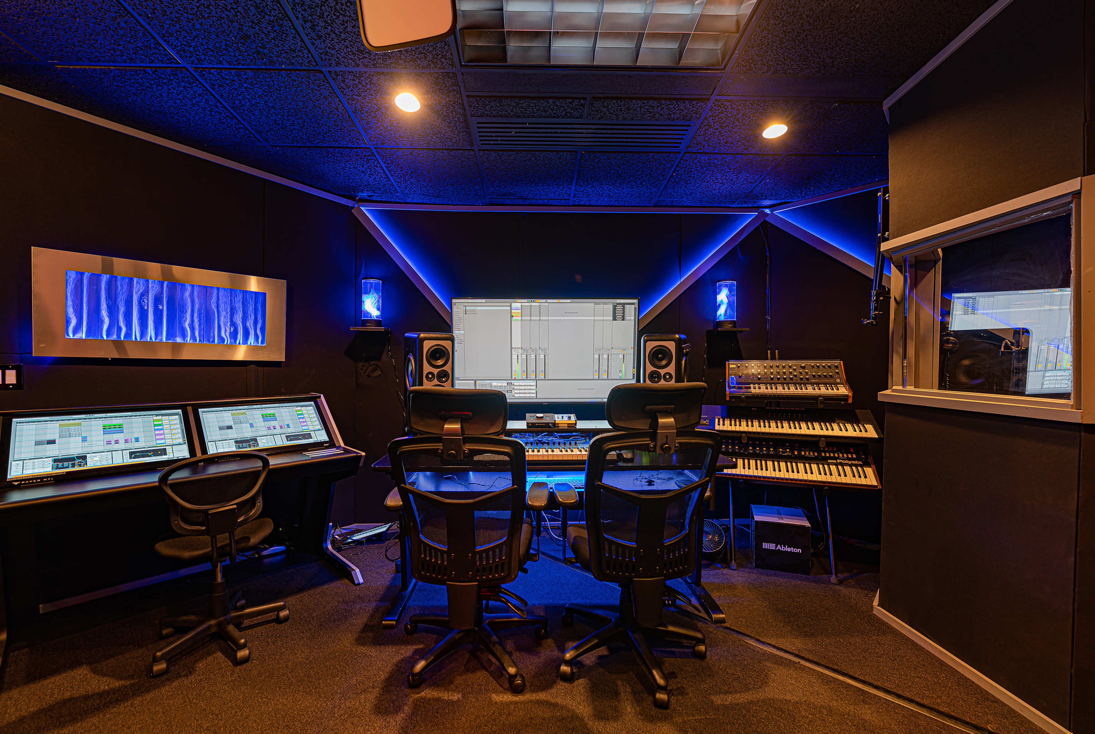 A modern music production studio with two computer workstations, synthesizers, and sound mixing equipment, illuminated by blue LED lighting.