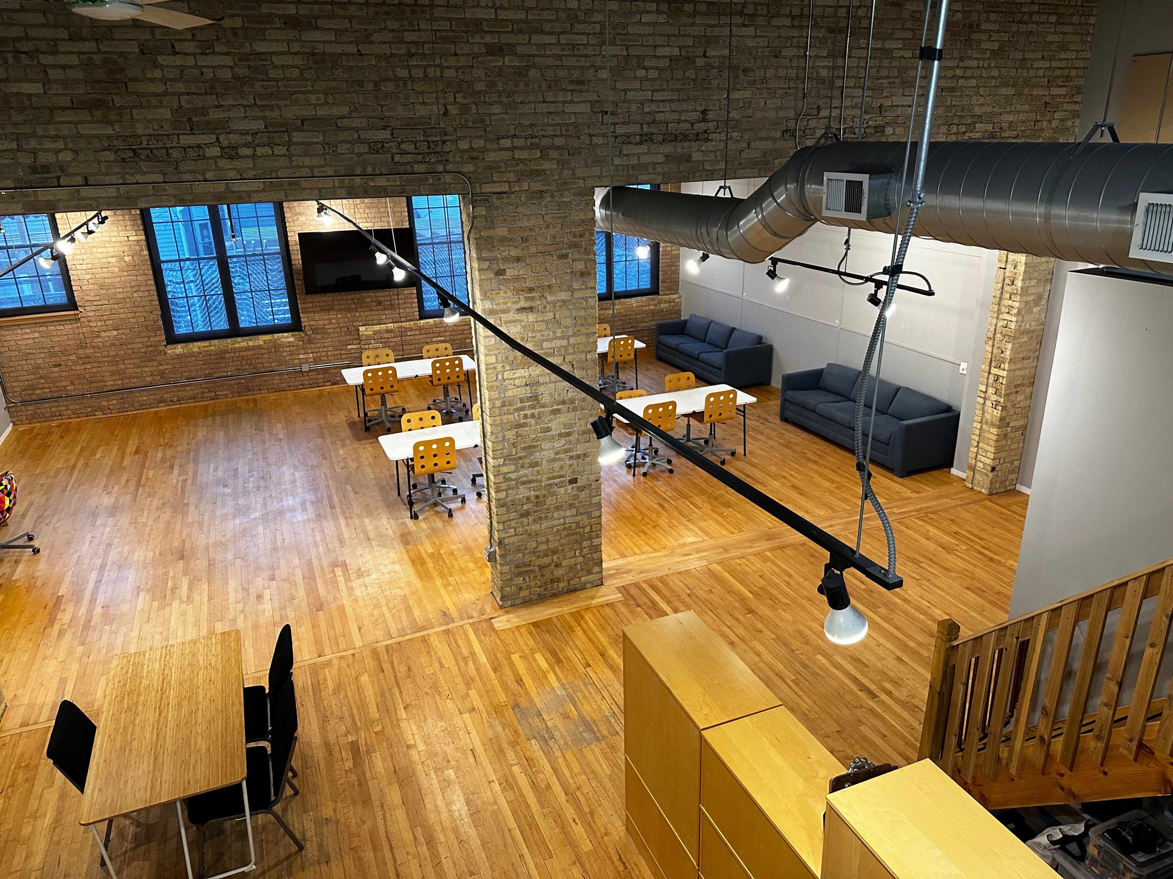 A spacious loft-style office with wooden floors, several desks with chairs, and a seating area with sofas against a brick wall.