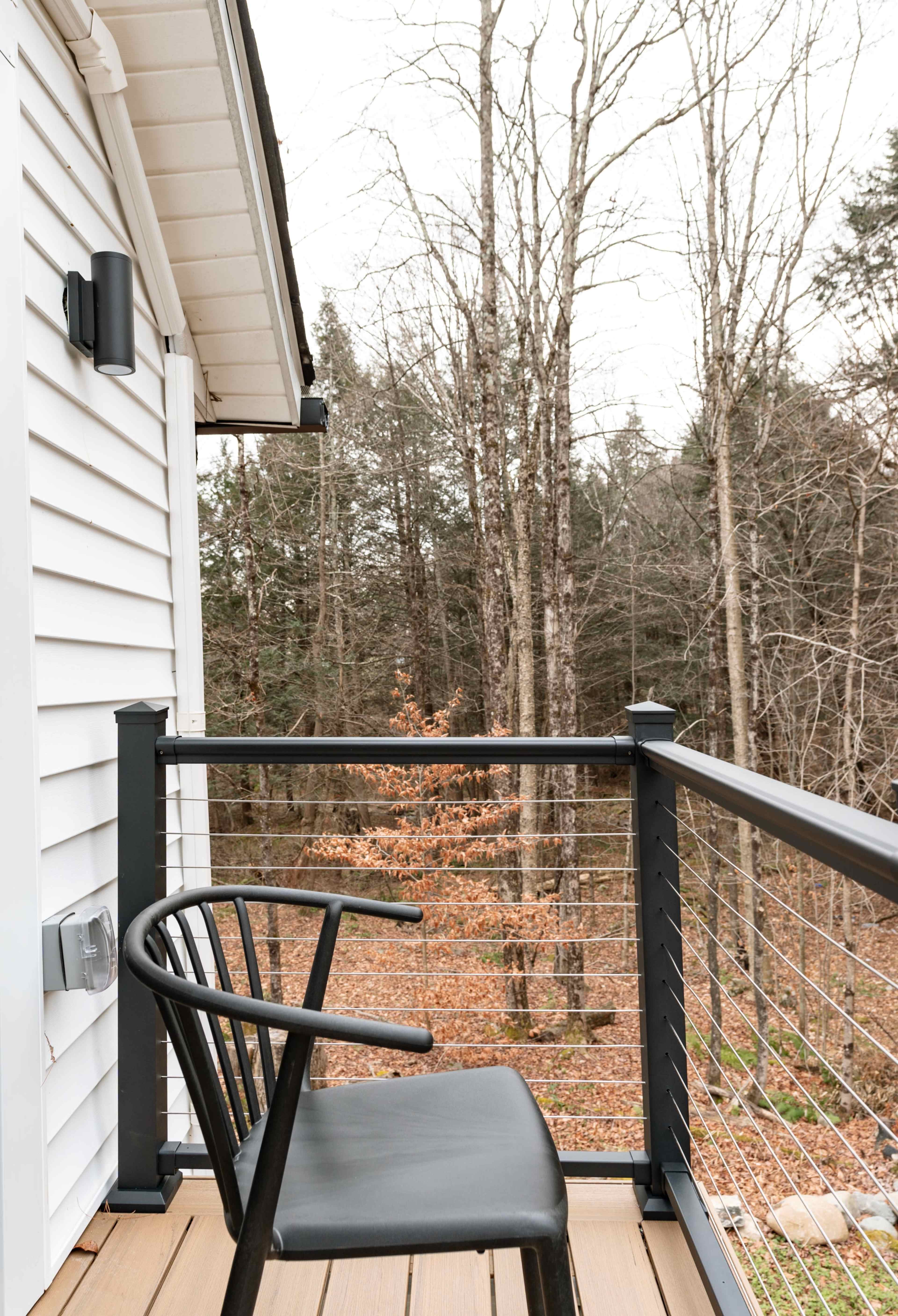 A black chair rests on a wooden balcony overlooking a wooded area with bare trees.