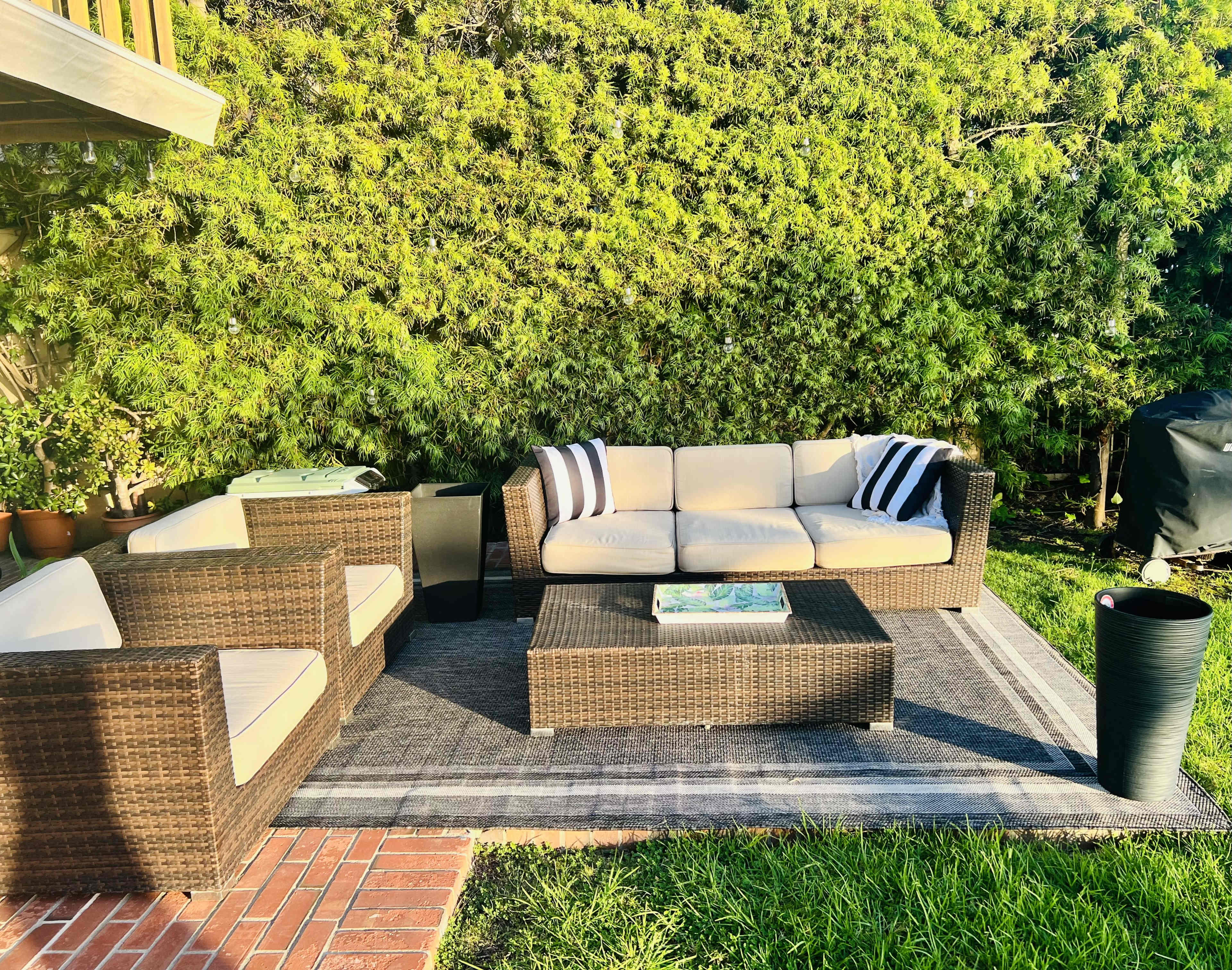 The image shows a cozy outdoor seating area with a wicker sofa and chairs arranged around a coffee table on a grassy surface, surrounded by tall greenery.