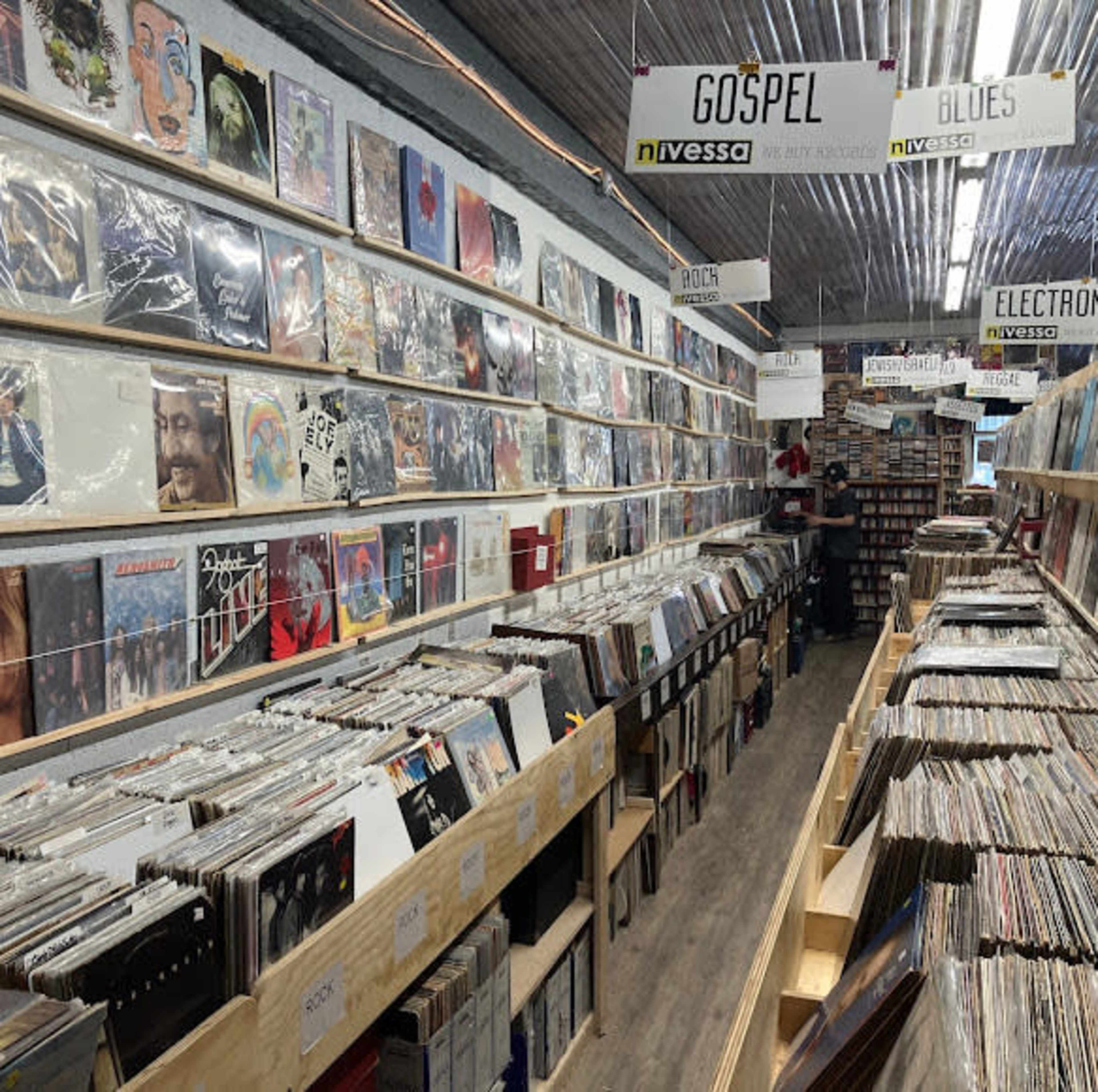 The image shows a record store interior with shelves lined with vinyl records organized by genre.