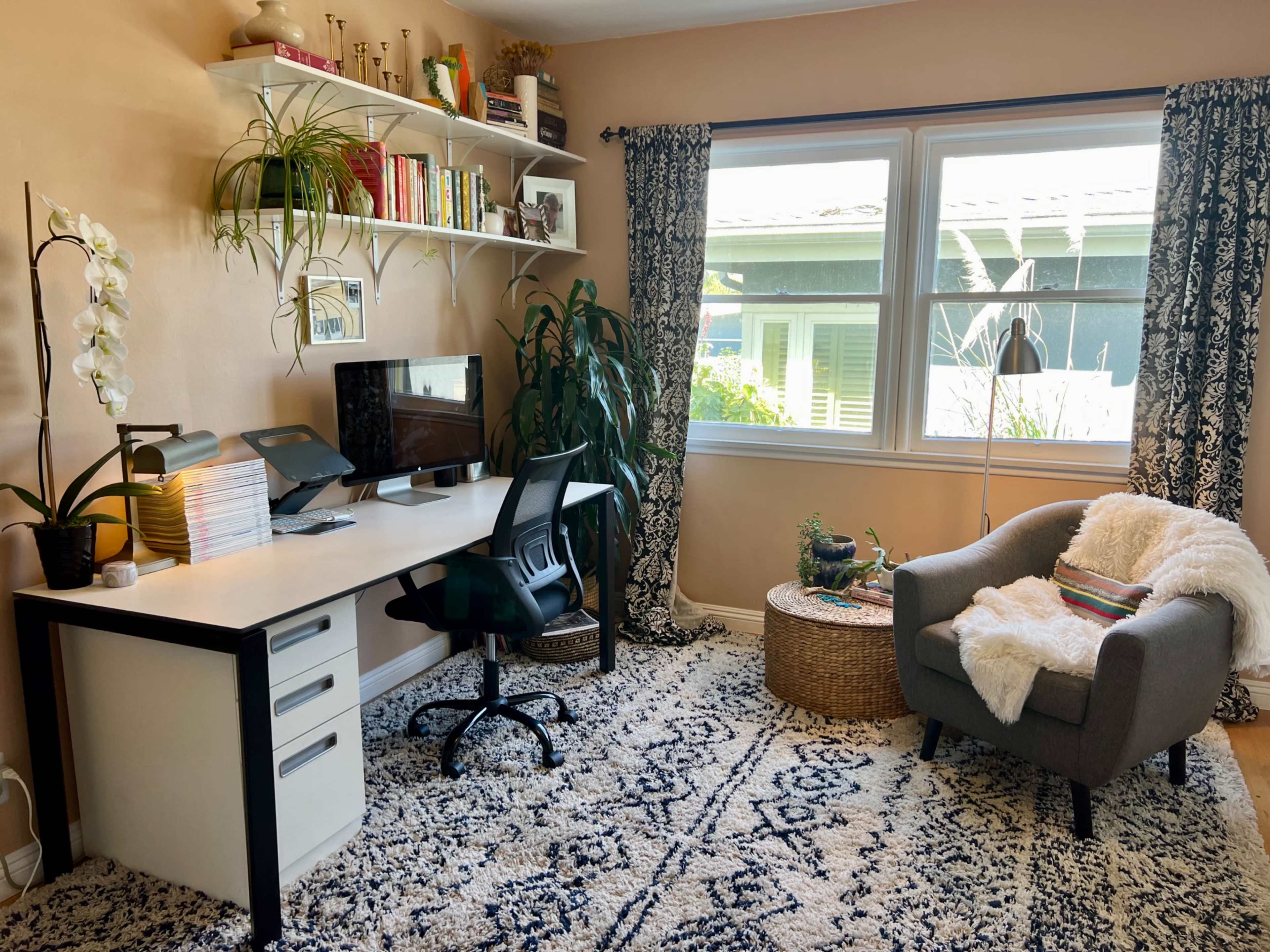 An organized home office features a desk with a computer, shelves filled with books, a cozy armchair, and plants near a window.