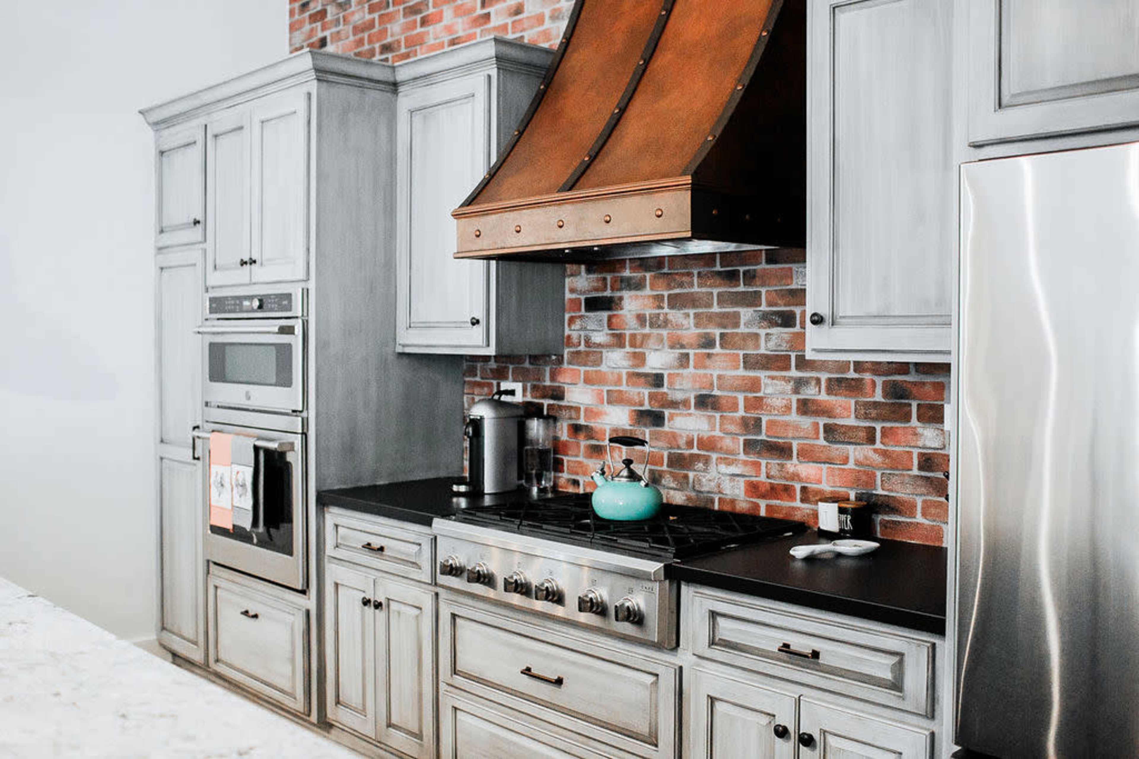 A modern kitchen with gray cabinetry, a brick backsplash, and a stainless steel appliance setup, including a stovetop and a range hood.