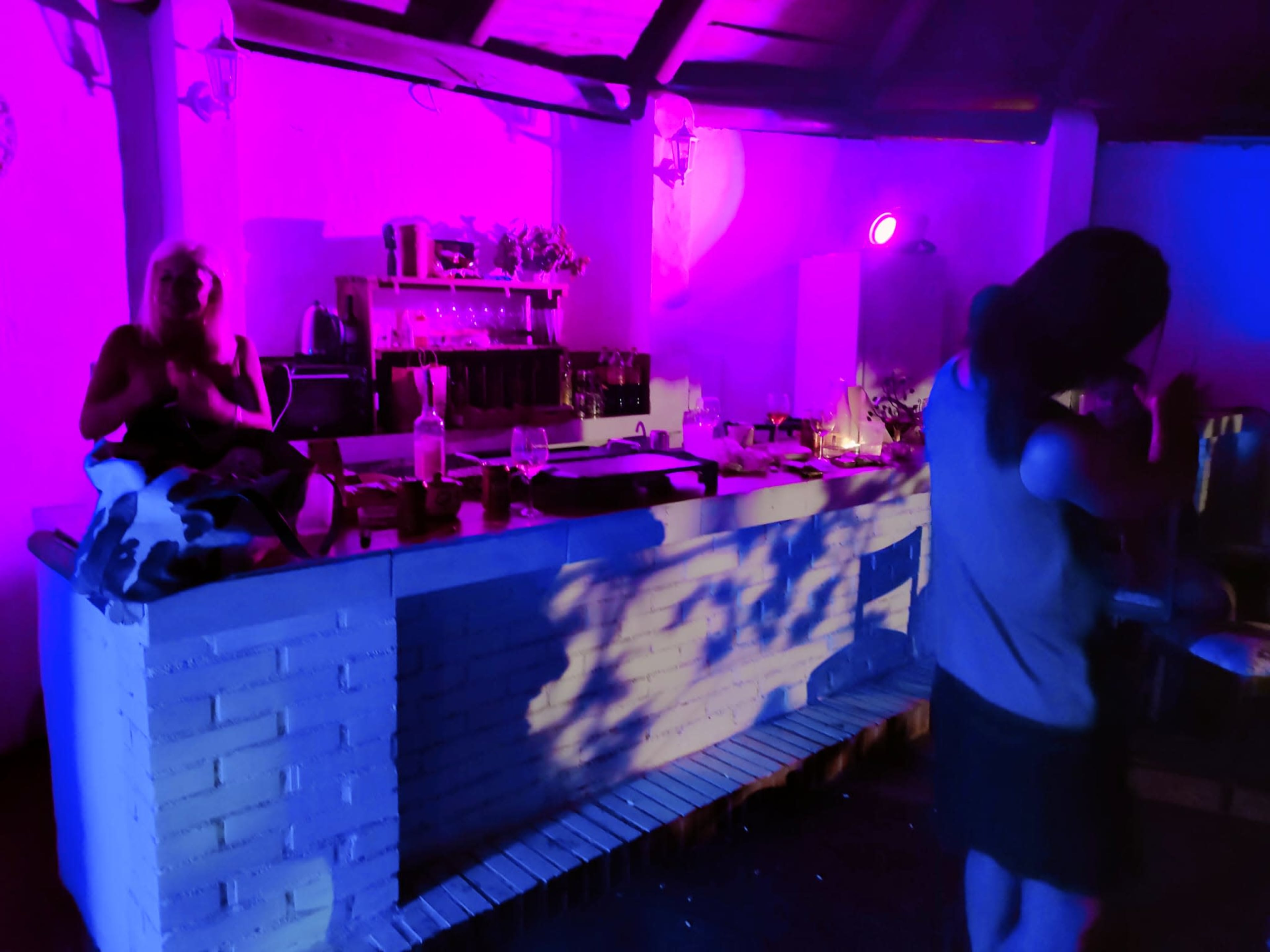 A dimly lit kitchen with purple and blue lighting, featuring a counter with various items and two people, one seated and another standing.