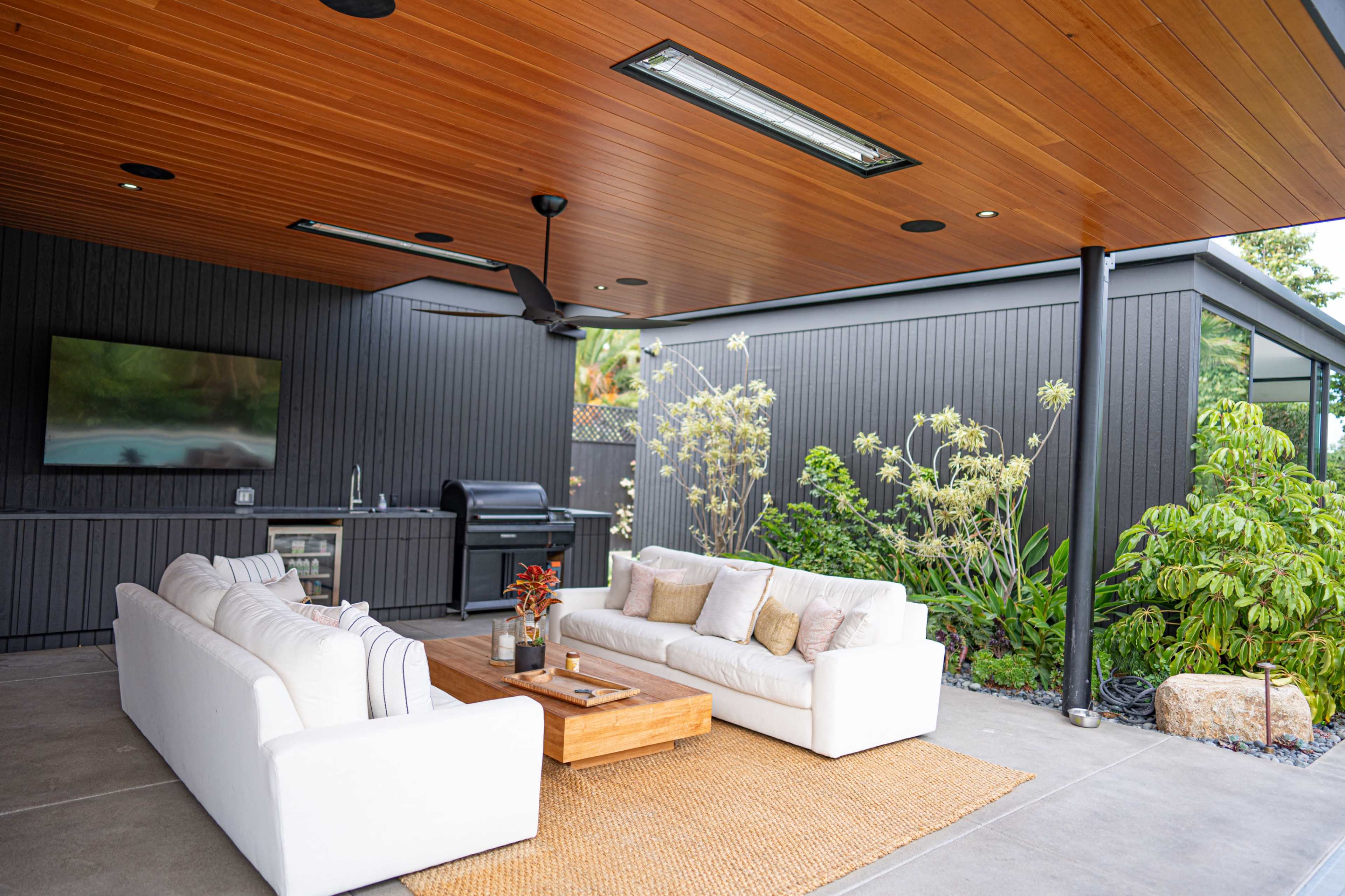 An outdoor living space with a spacious sofa, a wooden coffee table, a grill in the background, and plants surrounding the area, all under a wooden ceiling.