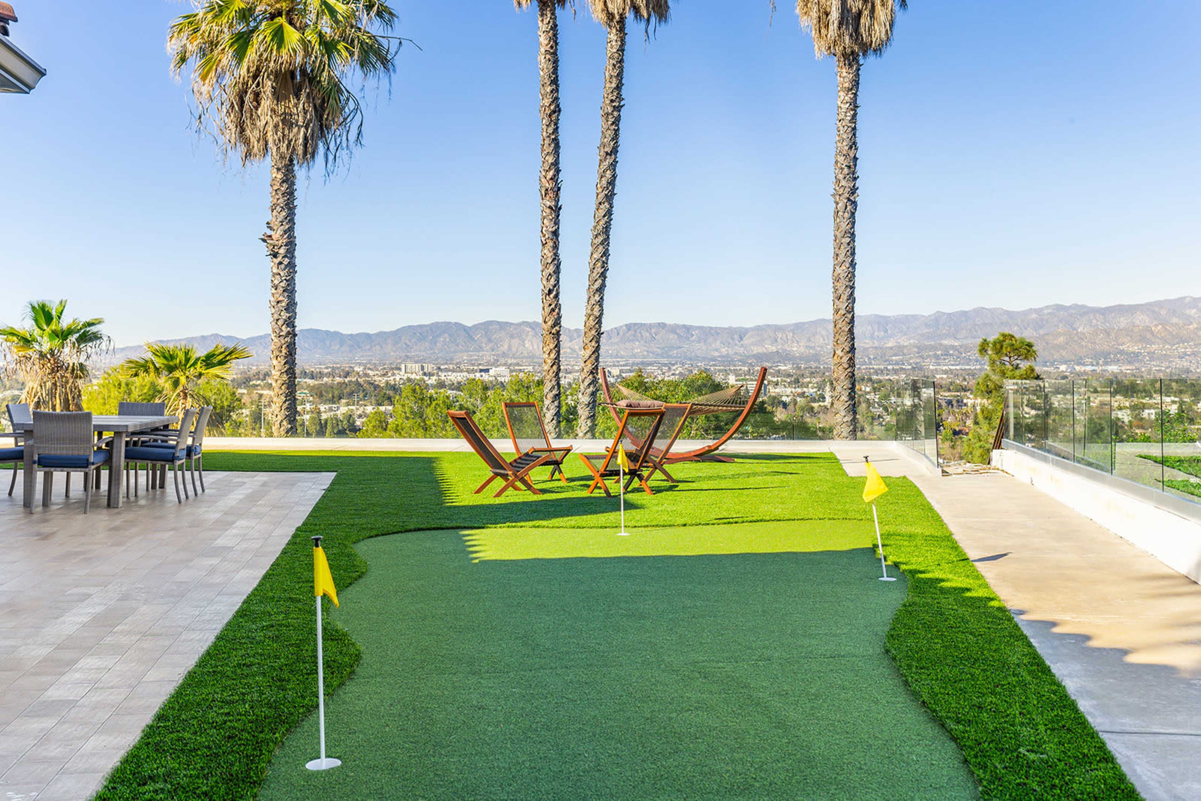 A well-maintained putting green with yellow flags, surrounded by palm trees and a view of the city in the background.