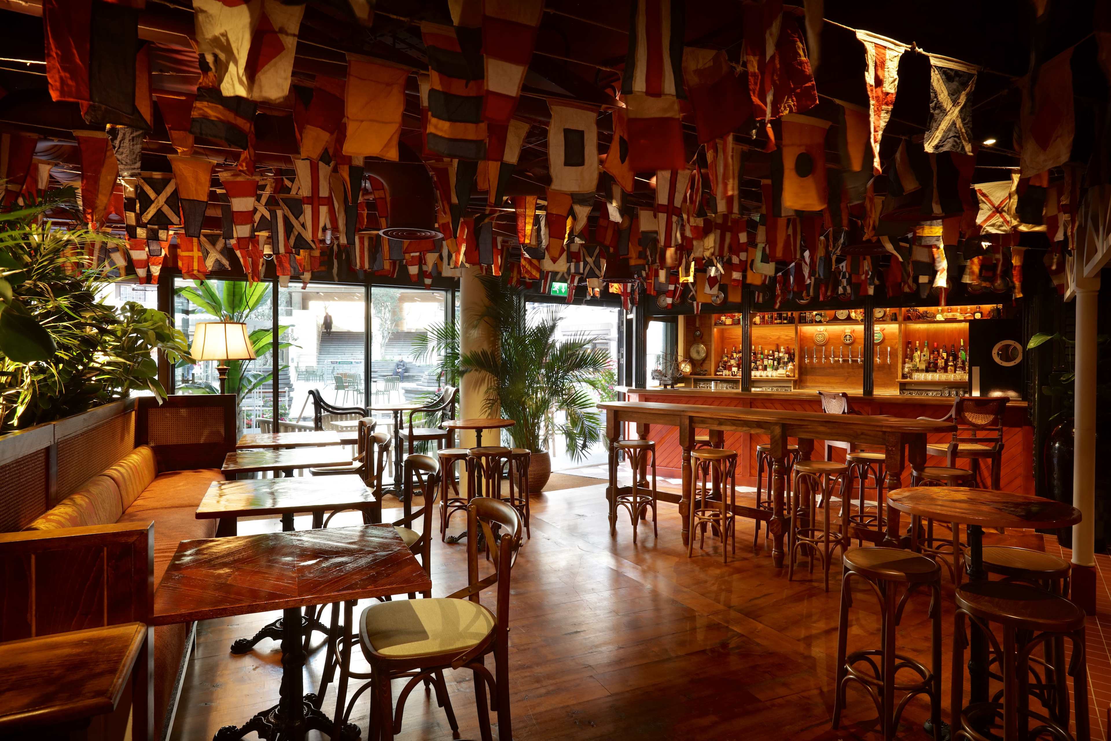 The image shows a bar interior decorated with various international flags hanging from the ceiling, with wooden tables and chairs arranged throughout the space.