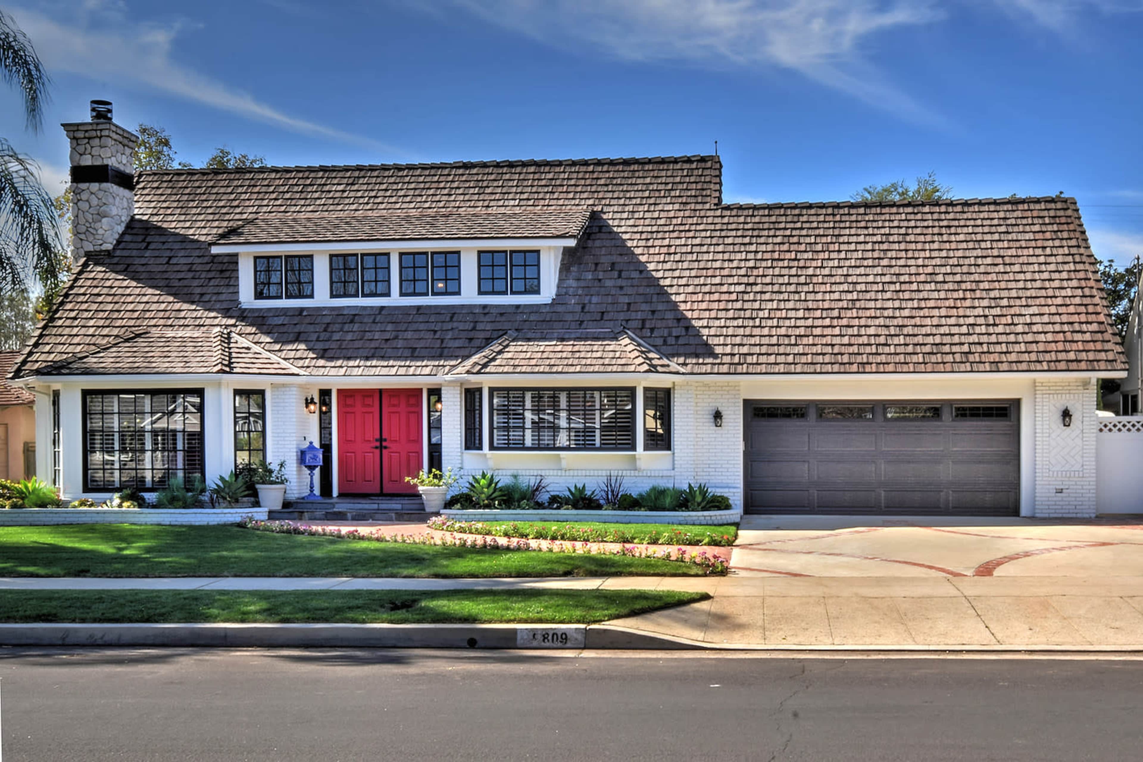 The image features a two-story house with a red door, white brick exterior, and a sloped roof, situated on a residential street.