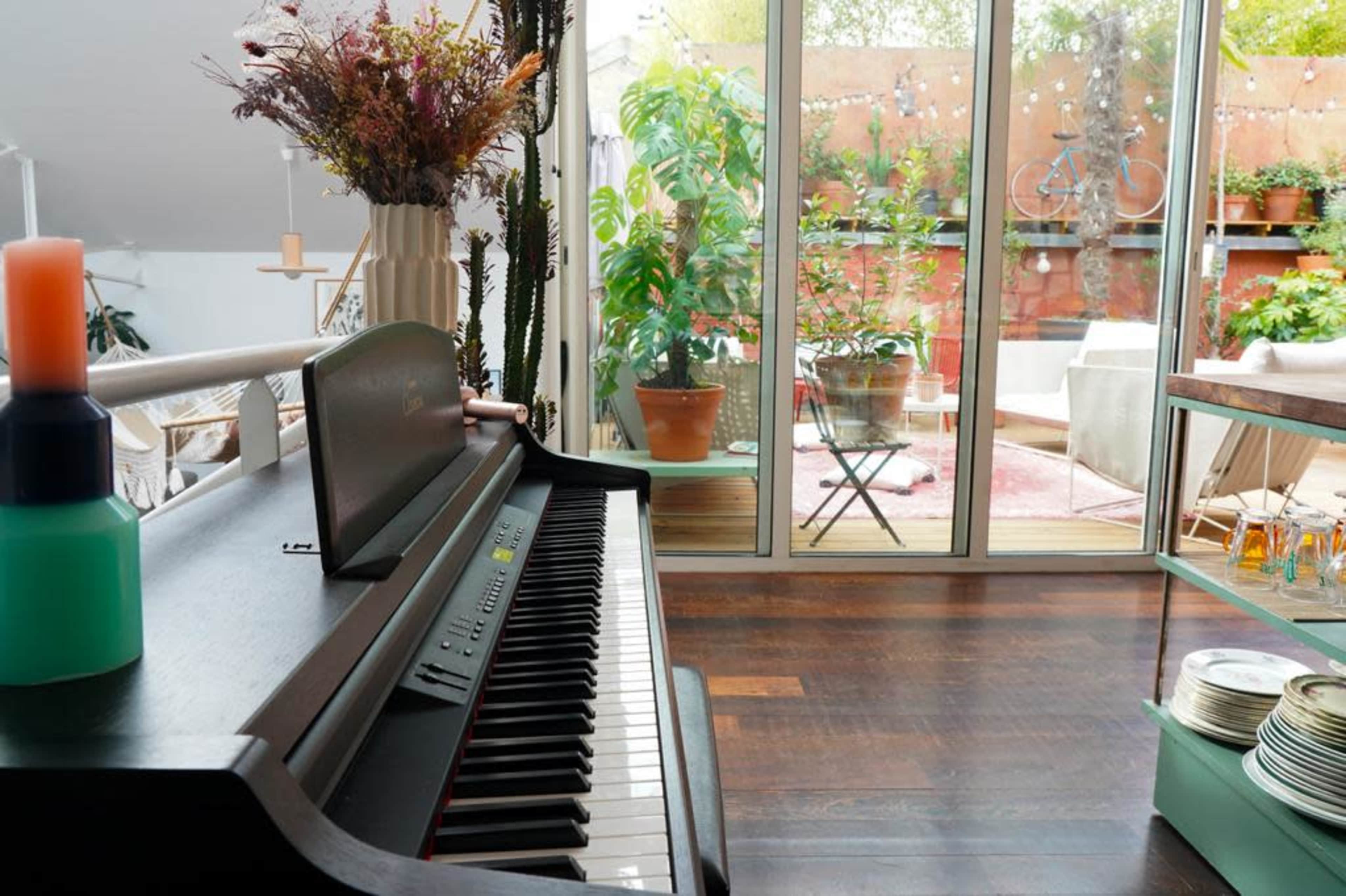A piano in the foreground, with a glass door leading to a patio filled with plants and outdoor furniture in the background.