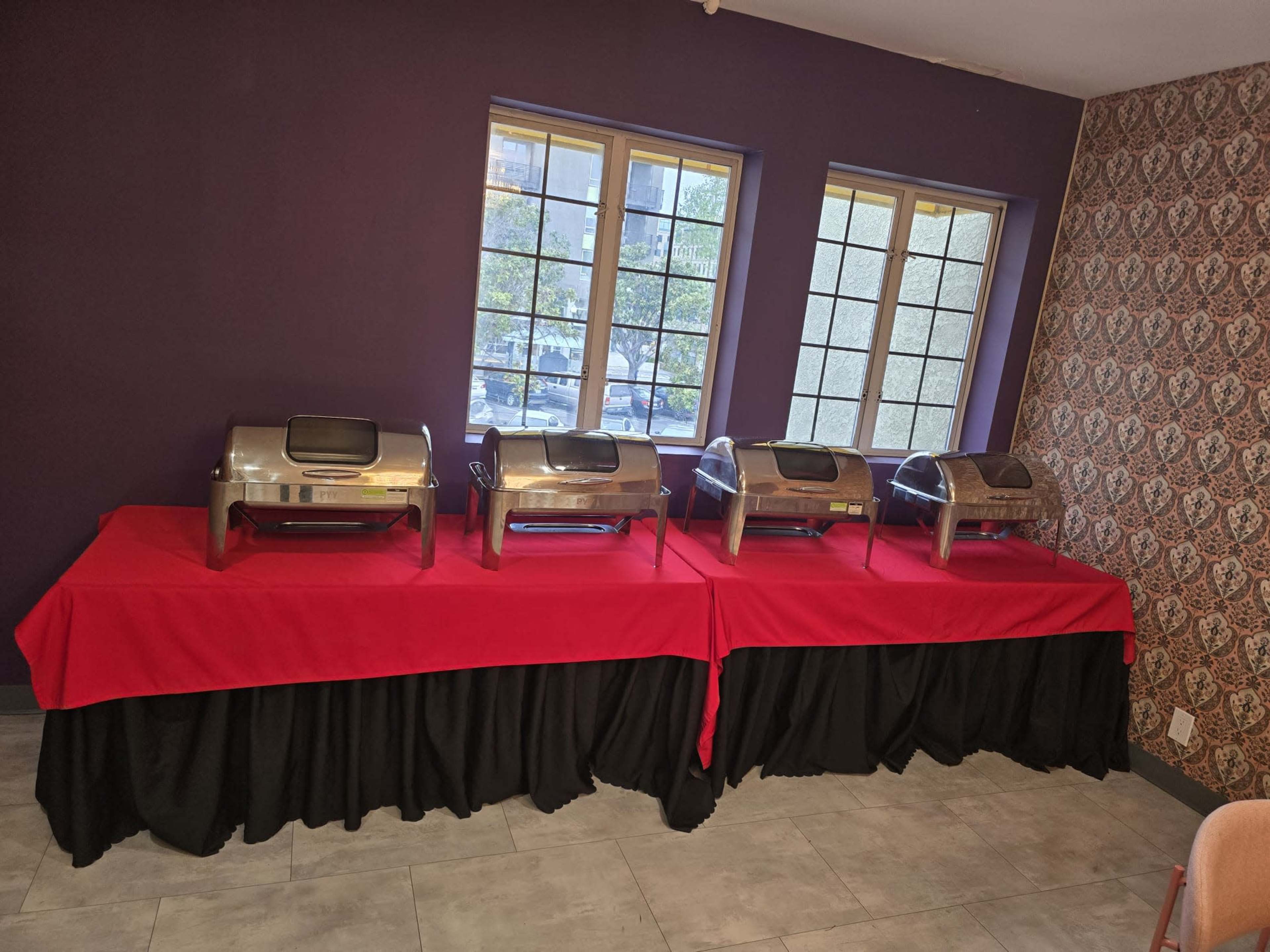 The image shows four chafing dishes arranged in a row on a table covered with a red cloth, positioned in front of two windows.