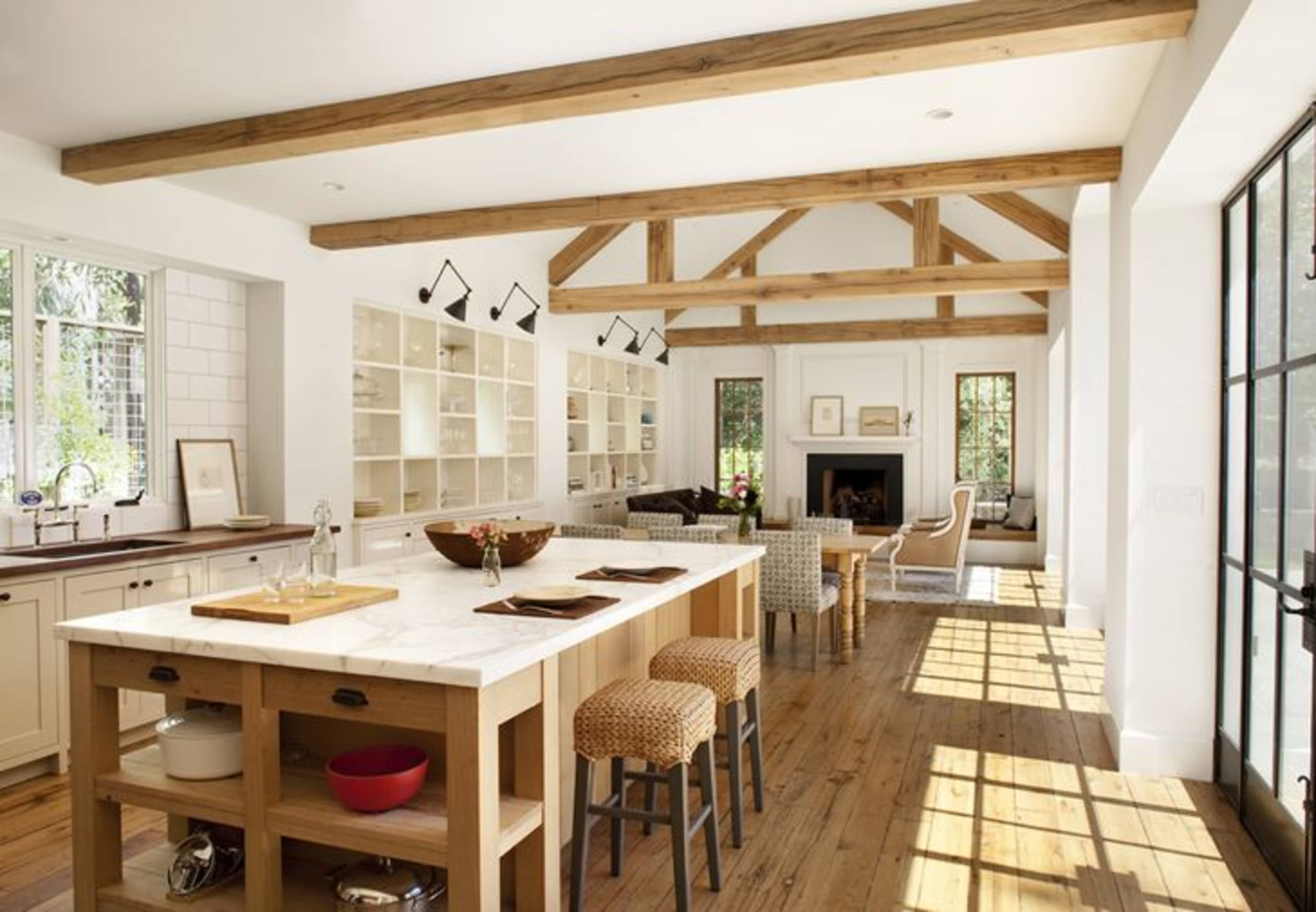 The spacious kitchen features a large marble island, wooden beams overhead, and ample natural light from large windows.