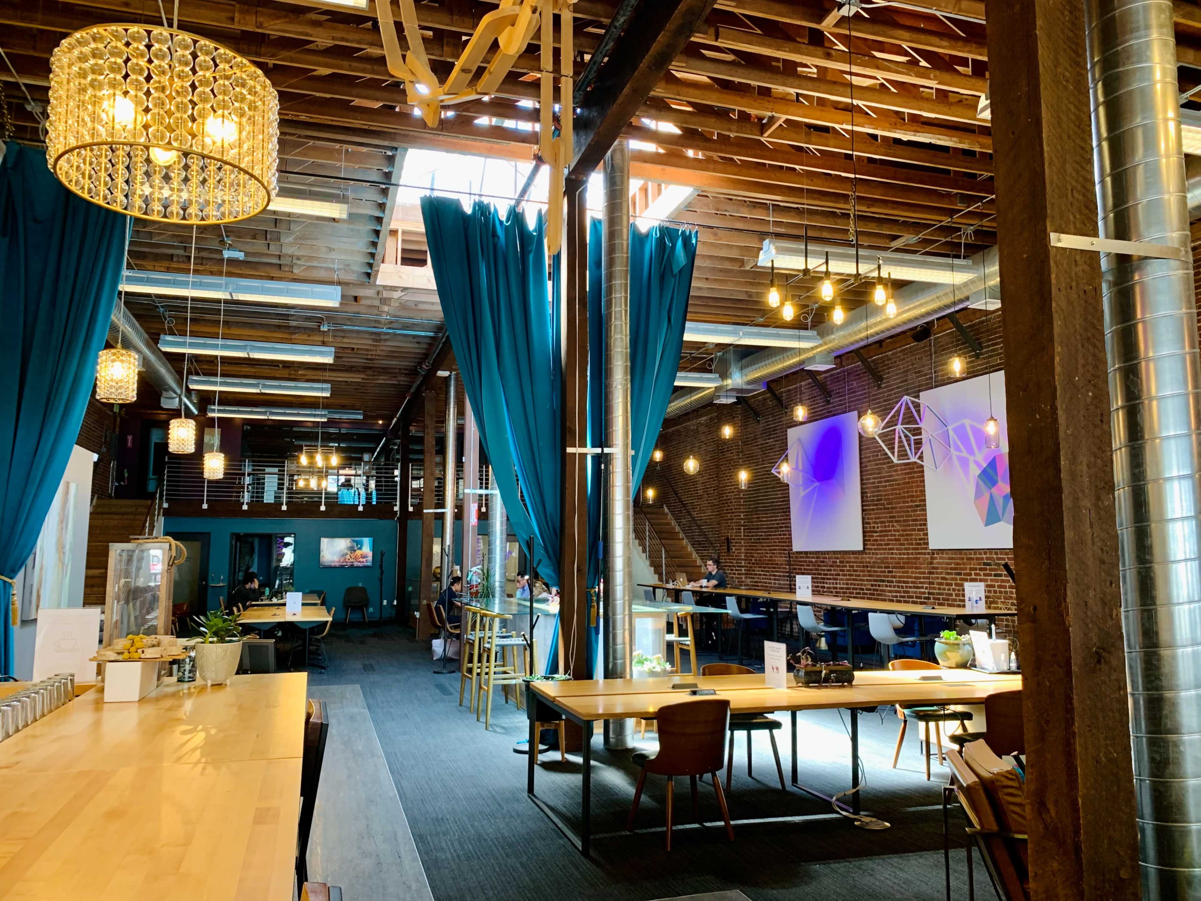 A spacious interior of a modern venue featuring high wooden ceilings, large blue curtains, and various seating areas with abstract artwork on the walls.