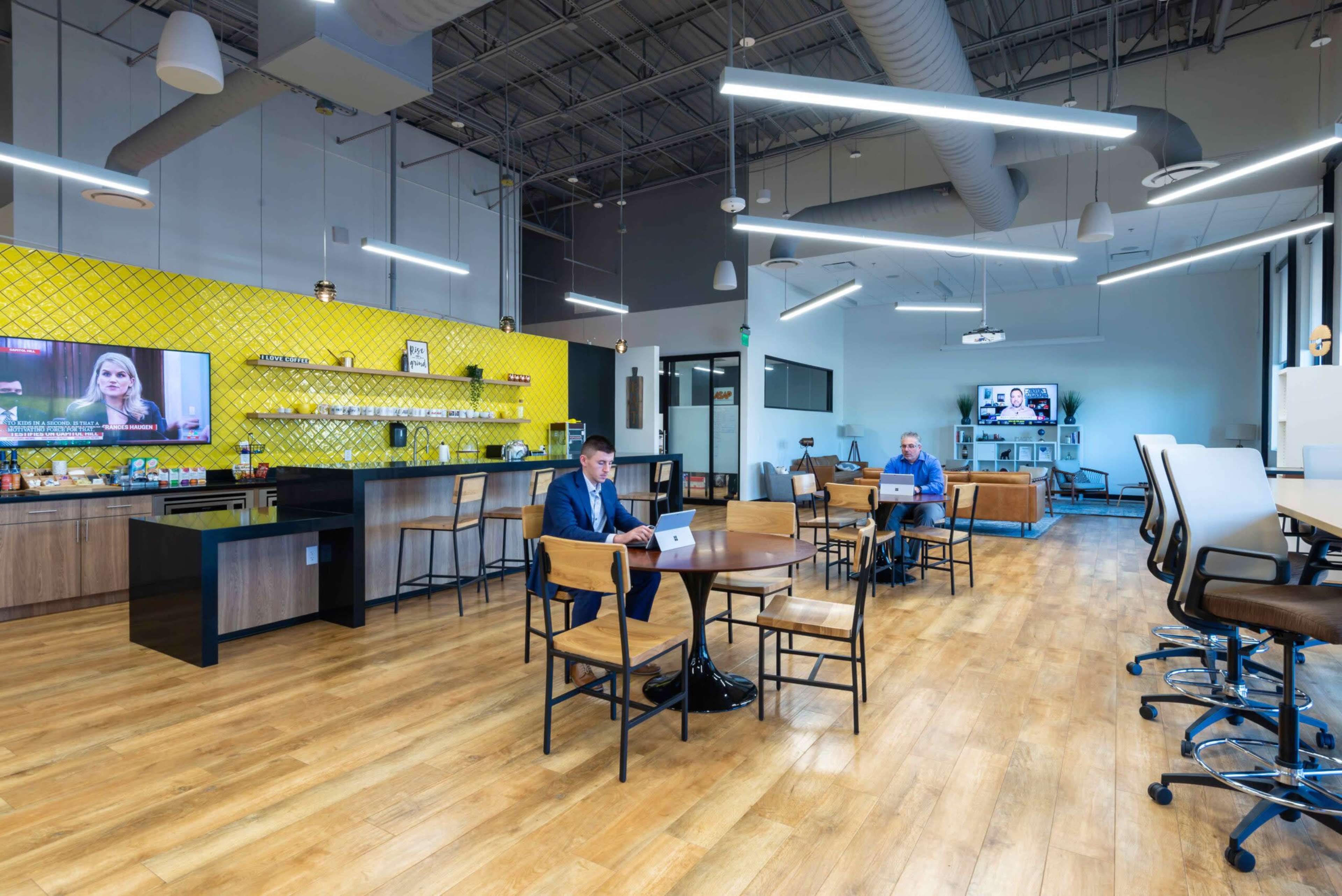 A modern office space features a large yellow accent wall, wooden flooring, and several workstations occupied by people using laptops.