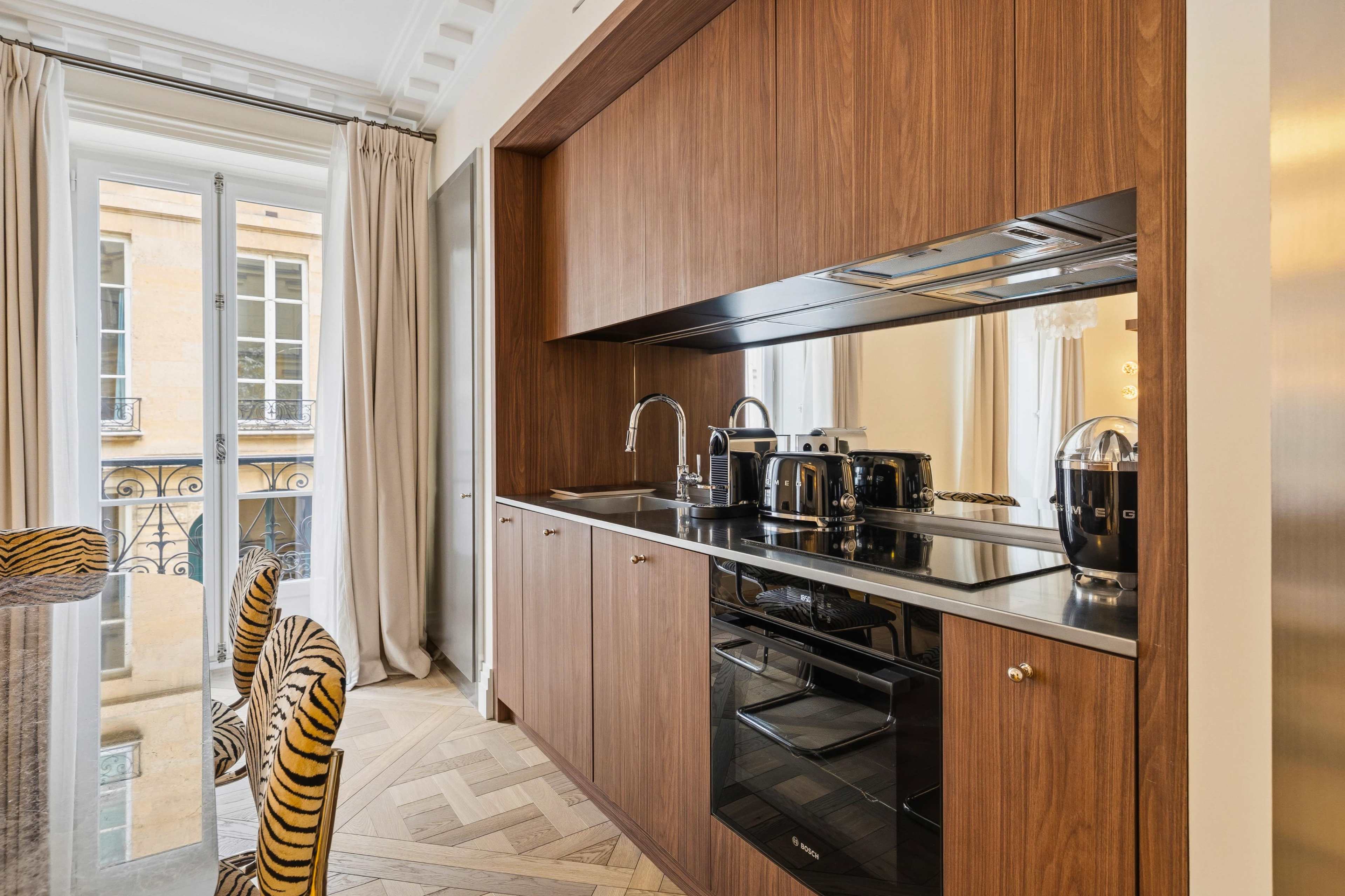 The image shows a modern kitchen with wooden cabinetry, stainless steel appliances, and large windows overlooking a city street.