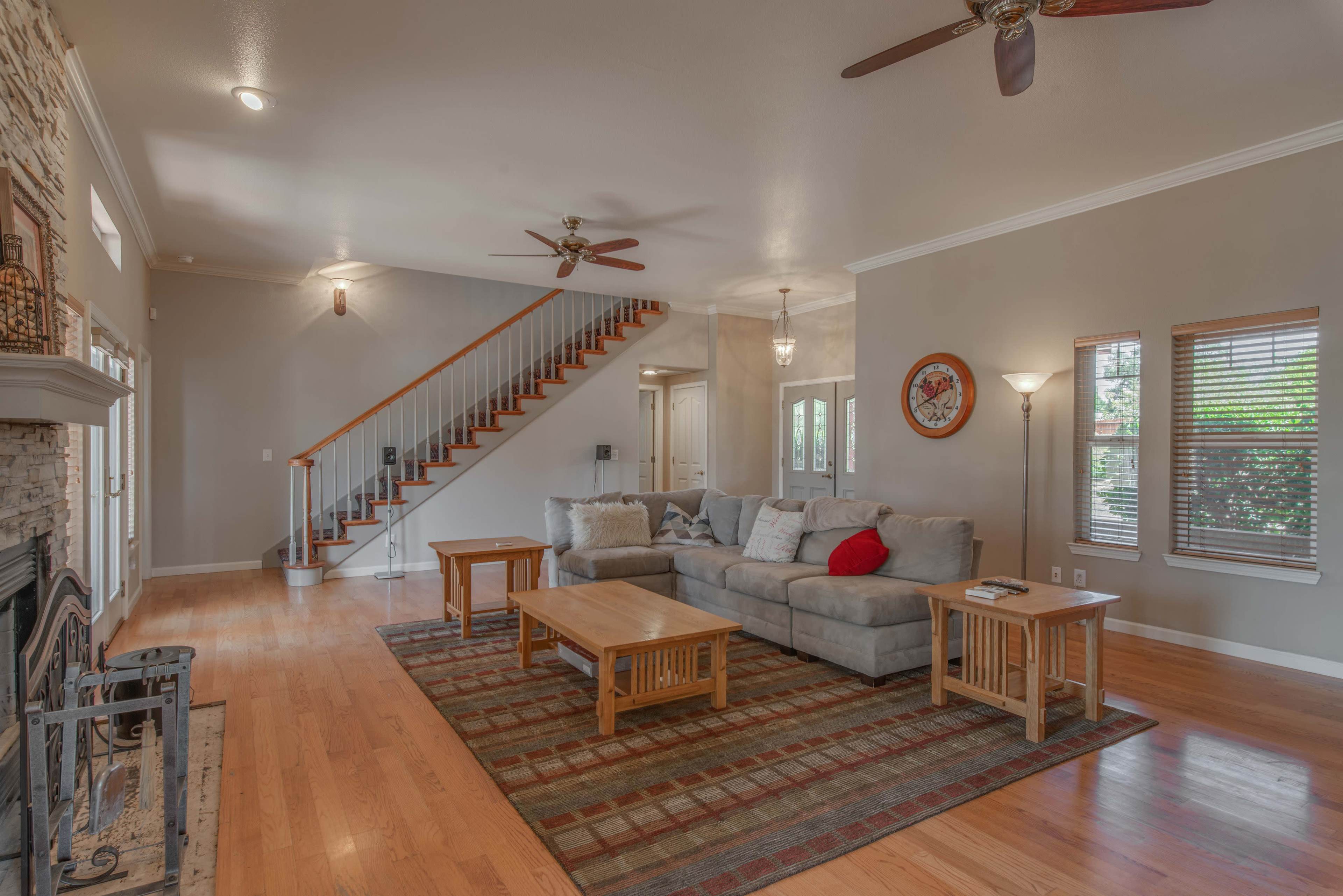 A spacious living room with a stone fireplace, a light gray couch, a wooden coffee table, and large windows, featuring a staircase in the background.