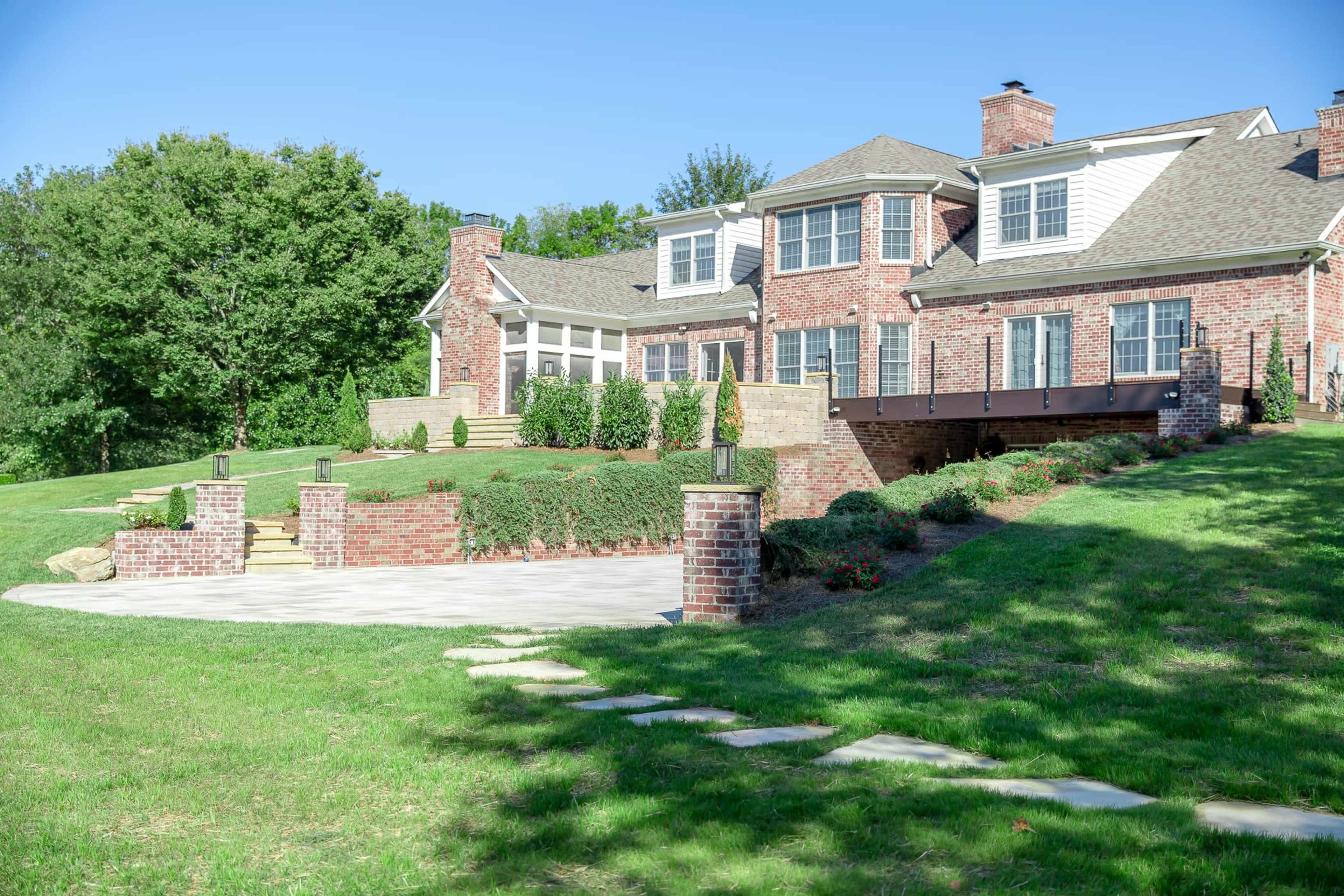 The image shows a brick house with a large patio, surrounded by a well-manicured lawn and landscaped gardens.