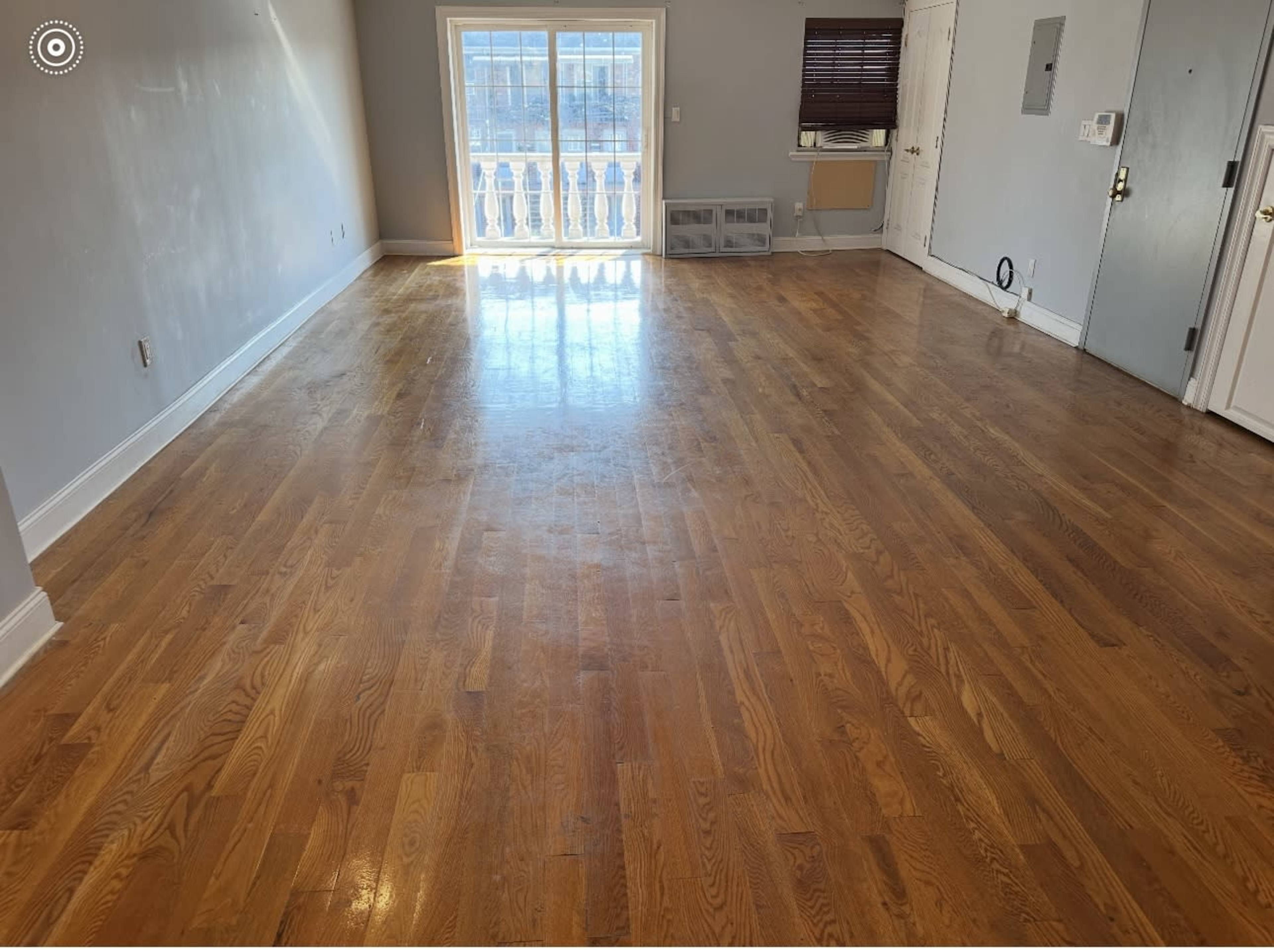 The image shows a large, empty room with hardwood flooring and a window overlooking a balcony.