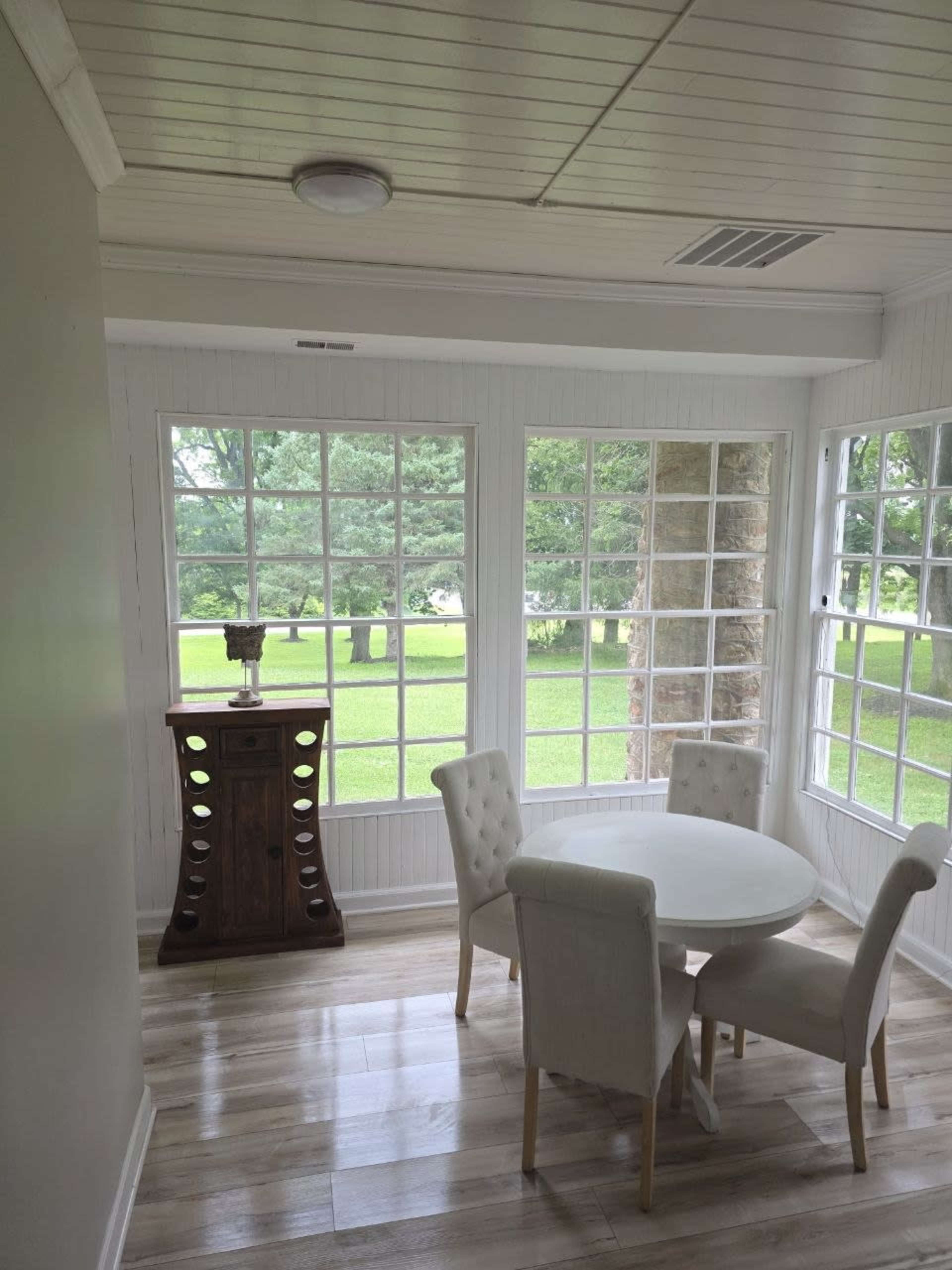 The image shows a sunlit dining area with a round white table and four upholstered chairs, surrounded by large windows that overlook a green outdoor space.