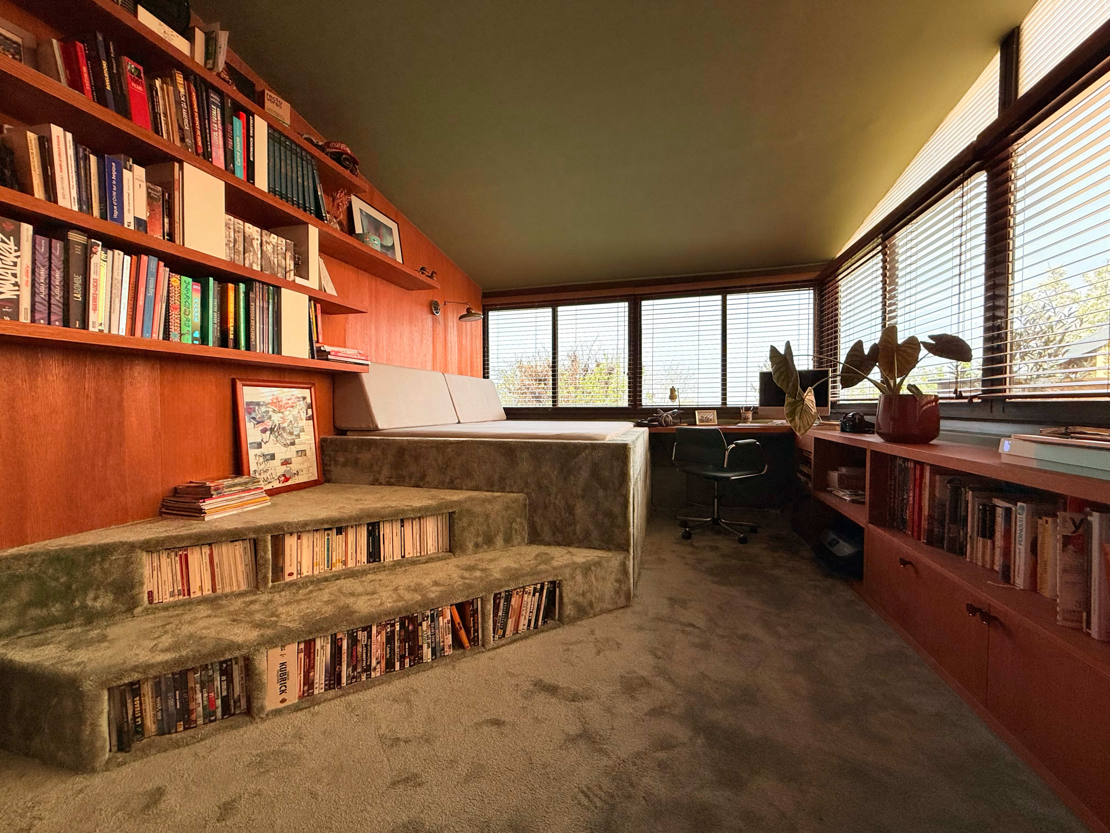 The image shows a cozy reading nook with a built-in bookshelf, a raised seating area, and large windows allowing natural light to fill the space.
