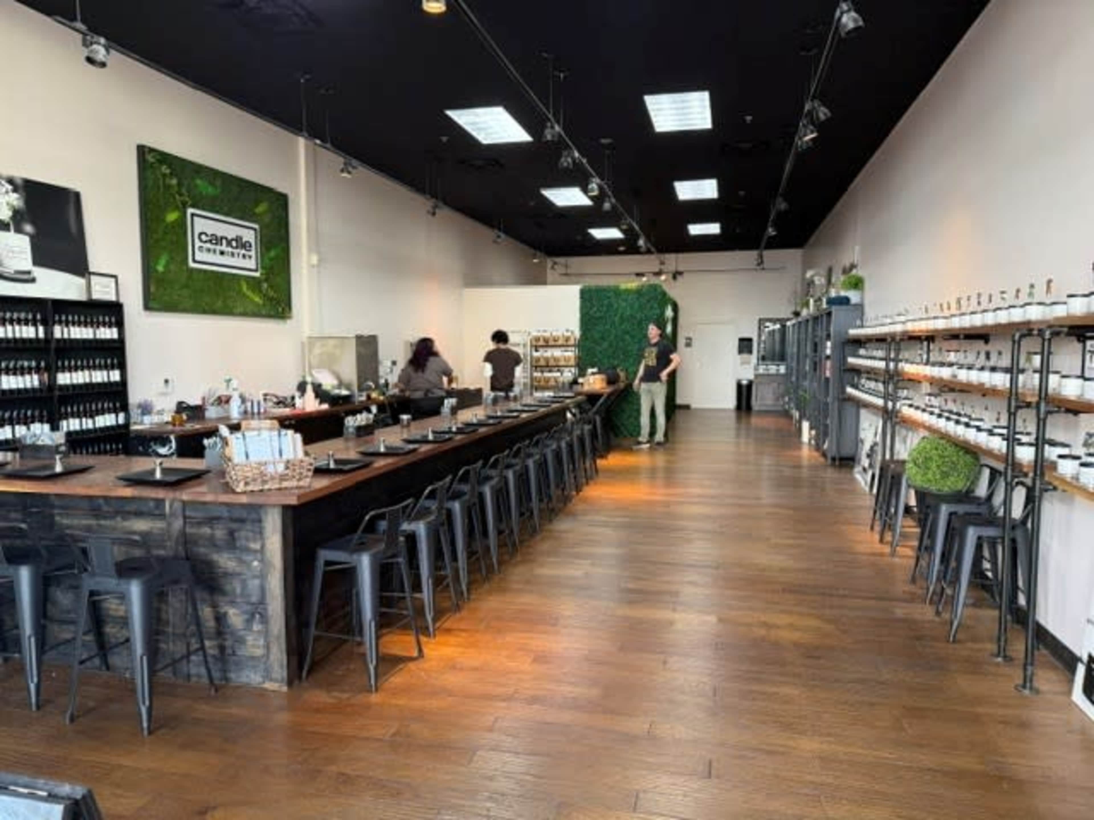 The image shows a spacious candle-making studio with a long wooden table and metal stools, lined with shelves displaying various candle supplies and greenery accents.