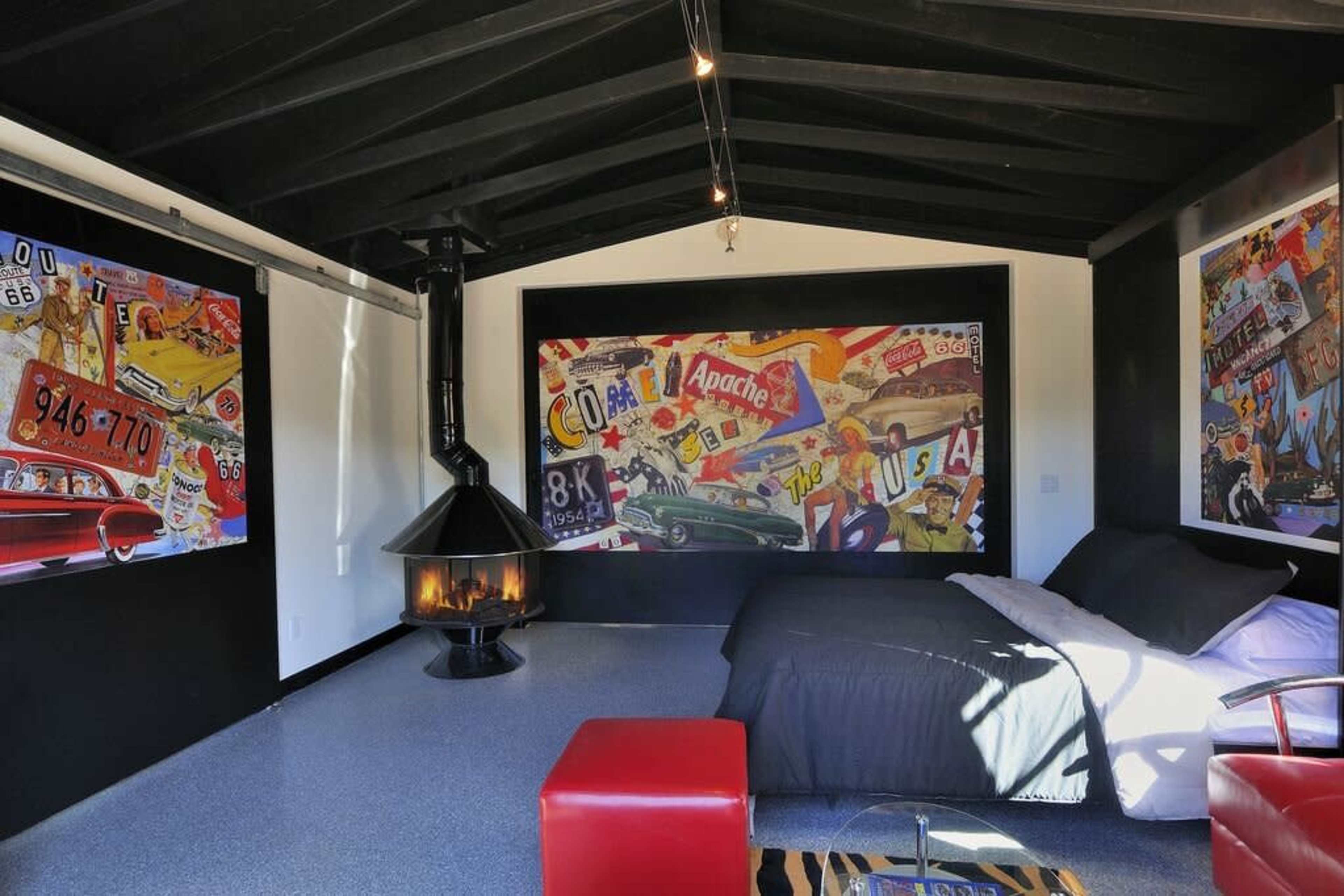 The image shows a modern bedroom featuring a bed, a red stool, and walls adorned with colorful vintage-style artwork, along with a black stove in the corner.